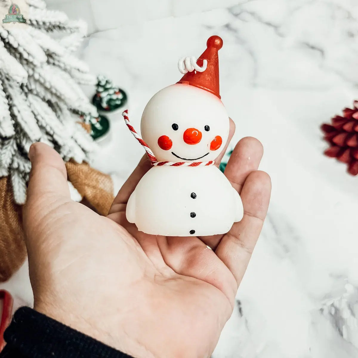 Someone holds a Snowman Candle featuring a red hat, red cheeks, orange nose, and striped scarf. Snowy decorations and a pine cone in the blurred background add cozy winter home decor vibes.
