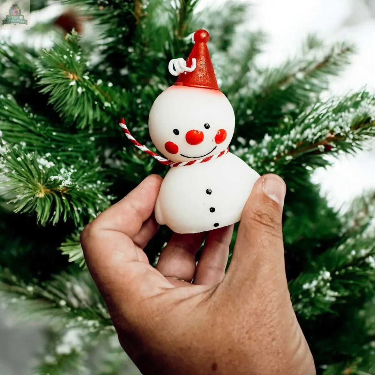 A hand holds the Snowman Candle with a red hat, red scarf, and black buttons in front of a snowy pine tree—perfect as a festive holiday gift or cheerful winter home decor.