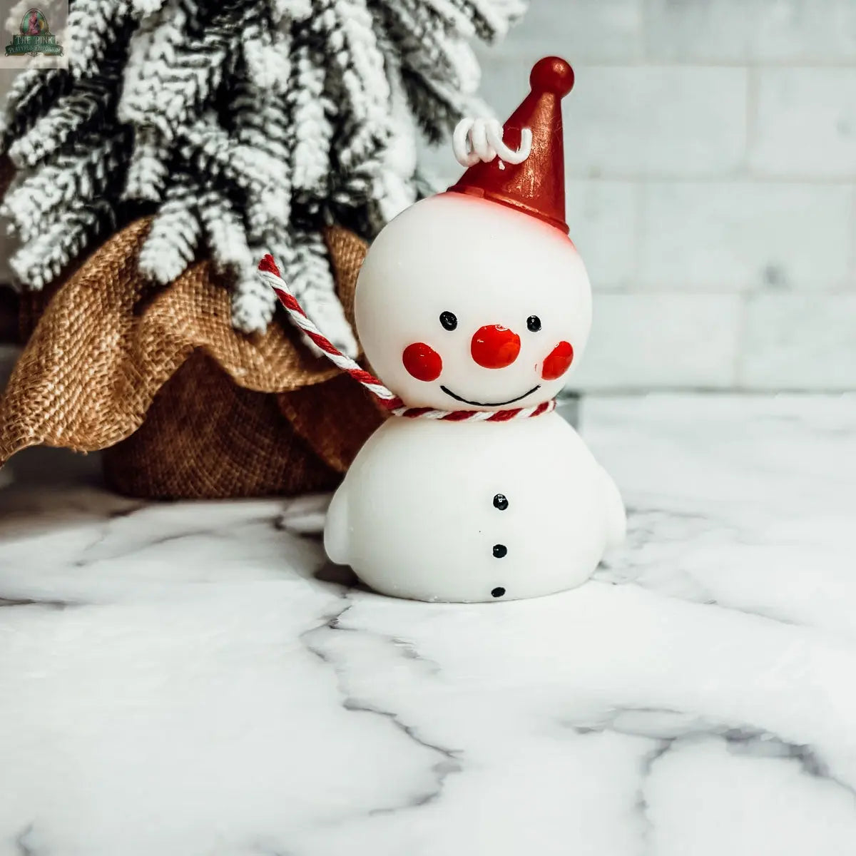 The Snowman Candle, featuring red cheeks, a red hat, and a striped scarf, sits on a marble surface—perfect for winter home decor. A frosted, burlap-wrapped tree accents the background.