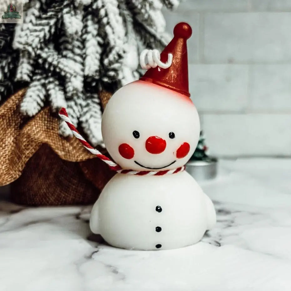 The Snowman Candle, featuring a red party hat, red nose, and striped scarf, sits on marble before a snow-dusted, burlap-wrapped Christmas tree—ideal as festive winter decor or a delightful holiday gift.