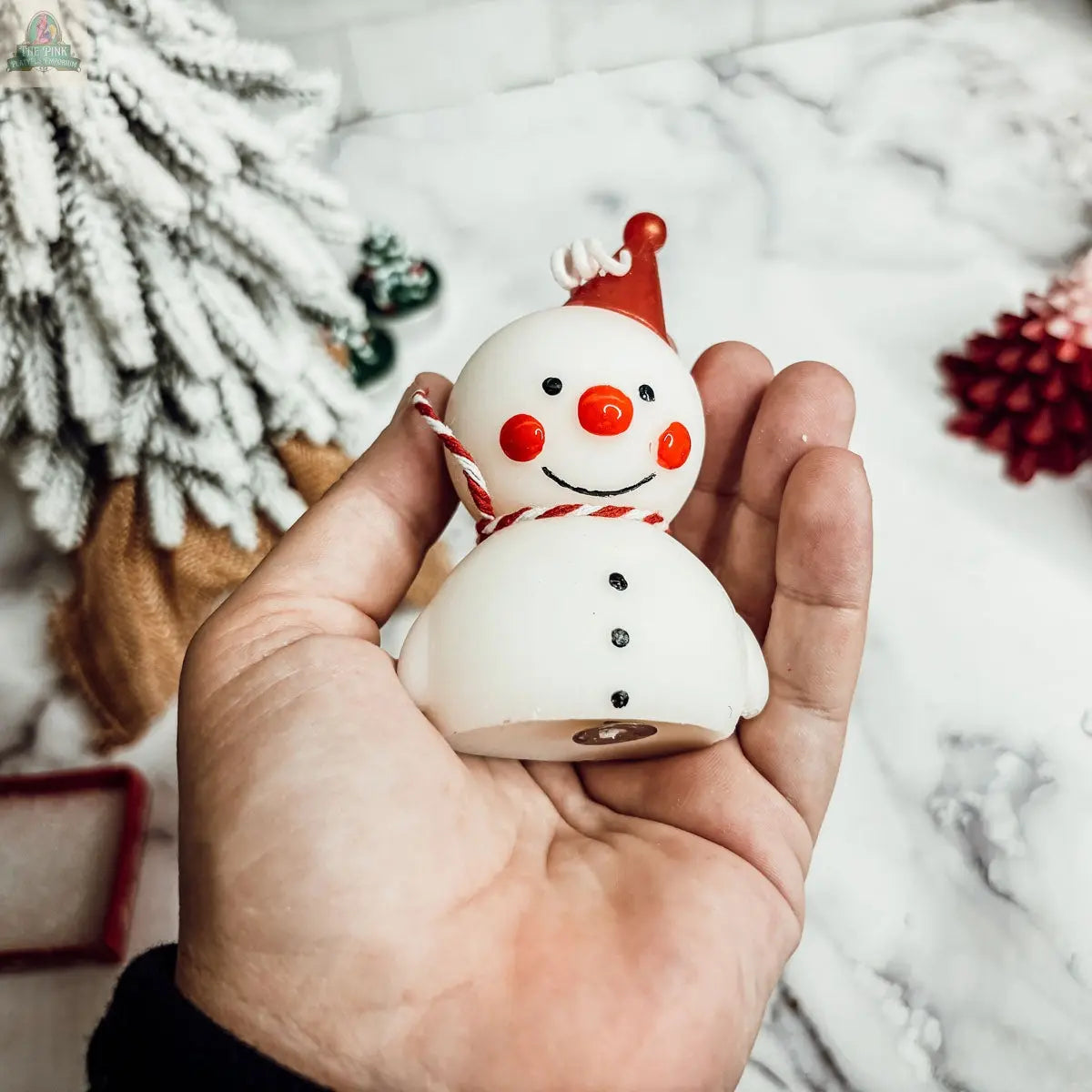 A hand holds the Snowman Candle, a small, round mini candle featuring a red hat, red cheeks, and a striped scarf. A snowy tree and red pinecone in the background add festive charm—ideal winter decor.
