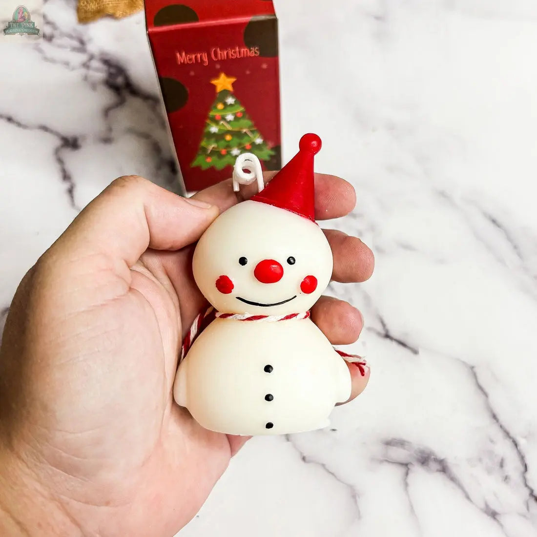 A hand holds a Snowman Candle adorned with a red hat and scarf—perfect winter home decor. Behind it, a festive red box with a Christmas tree and “Merry Christmas” message makes this an ideal holiday gift.