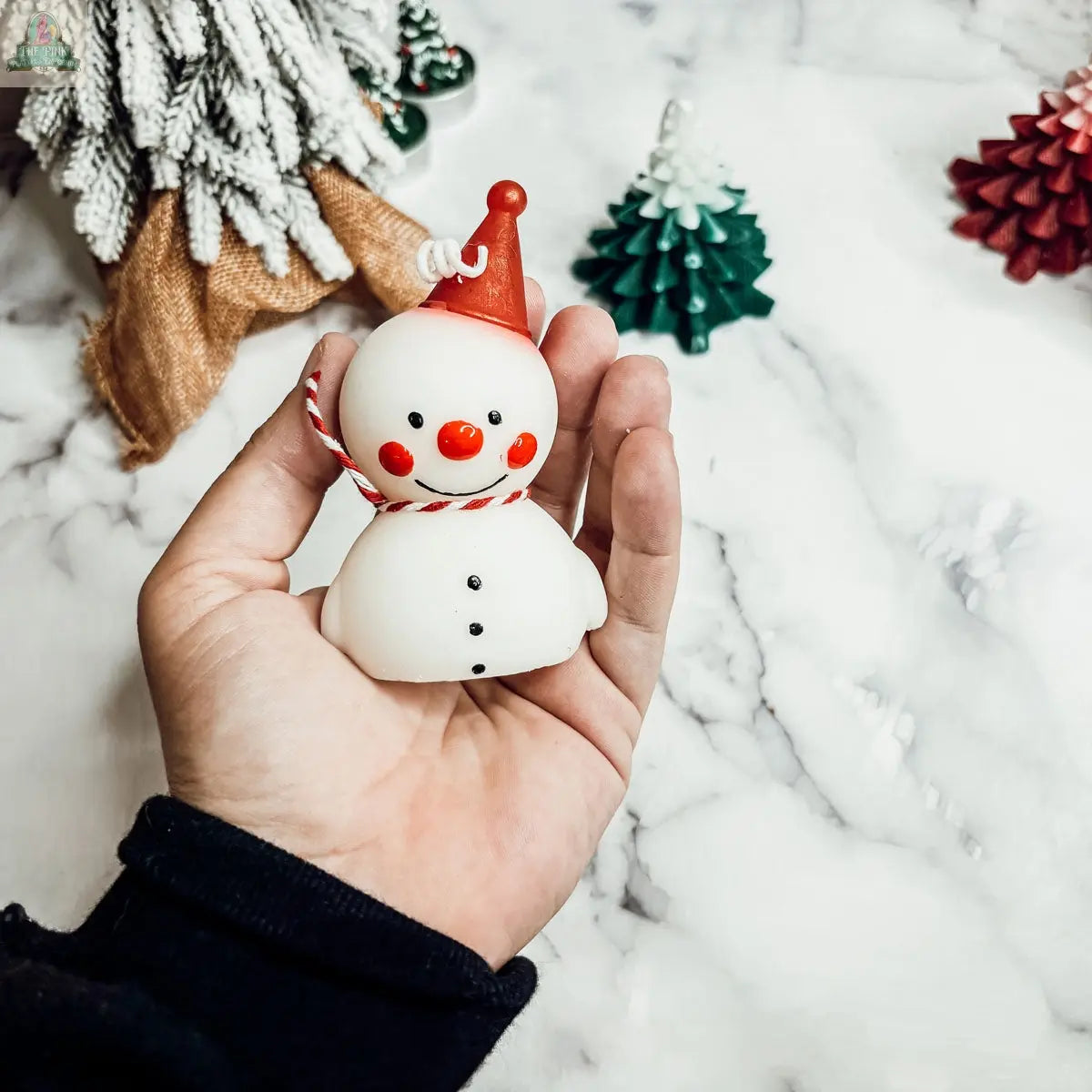 A hand holds the Snowman Candle, featuring a red Santa hat and a candy cane striped scarf. Festive green and red Christmas tree ornaments sit on a white marble surface in the background, ideal for winter home decor.