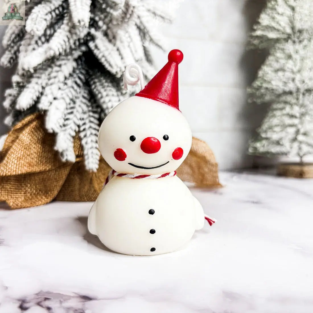 A cheerful Snowman Candle with a red hat, scarf, and buttons smiles on a marble surface—ideal for winter home décor or as a holiday gift—with snowy, burlap-wrapped trees in the background.