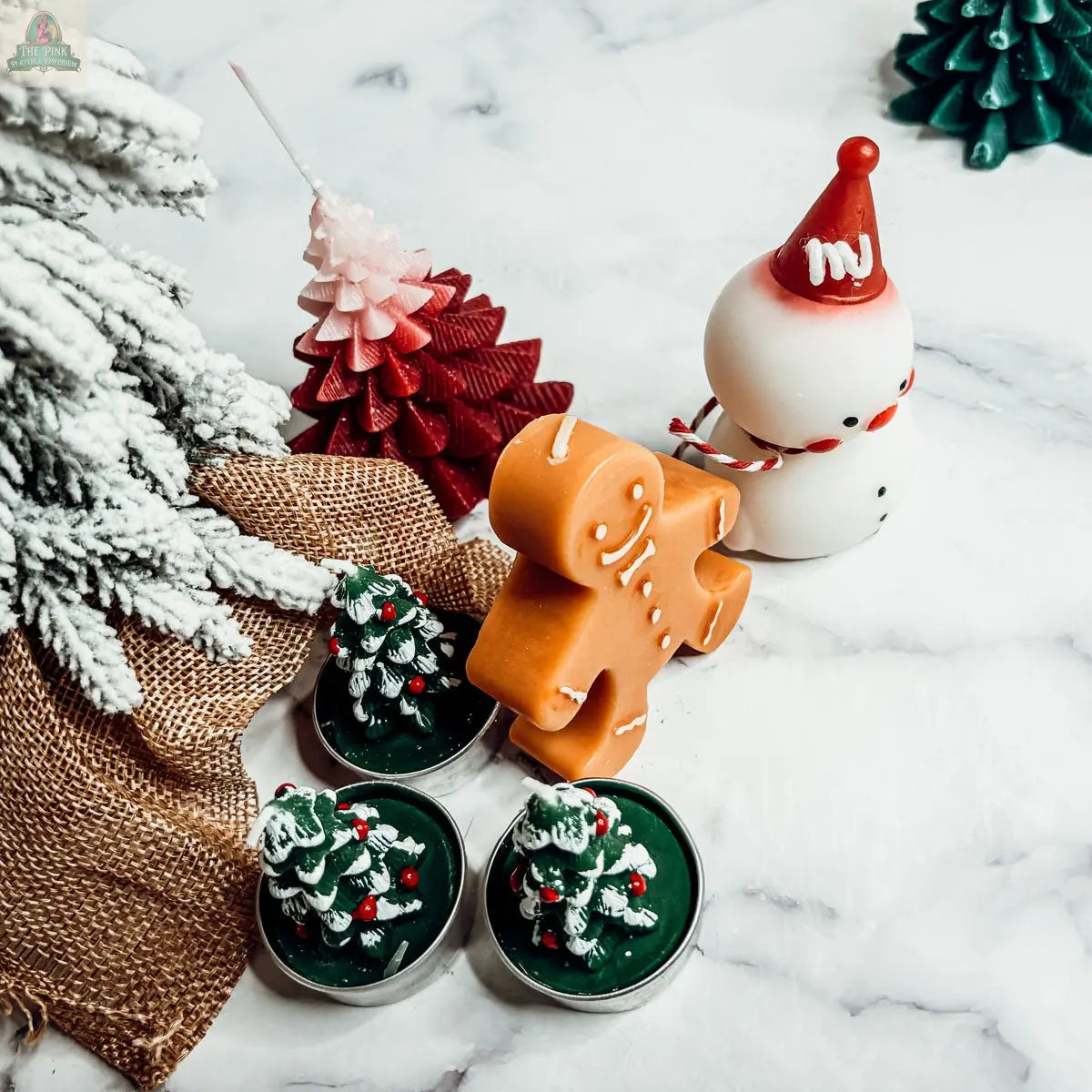 A festive scene featuring the Snowman Candle in a red hat, two green tree-shaped candles, and a pink Christmas tree candle decorates this white marble surface—ideal as winter home decor or a delightful holiday gift.