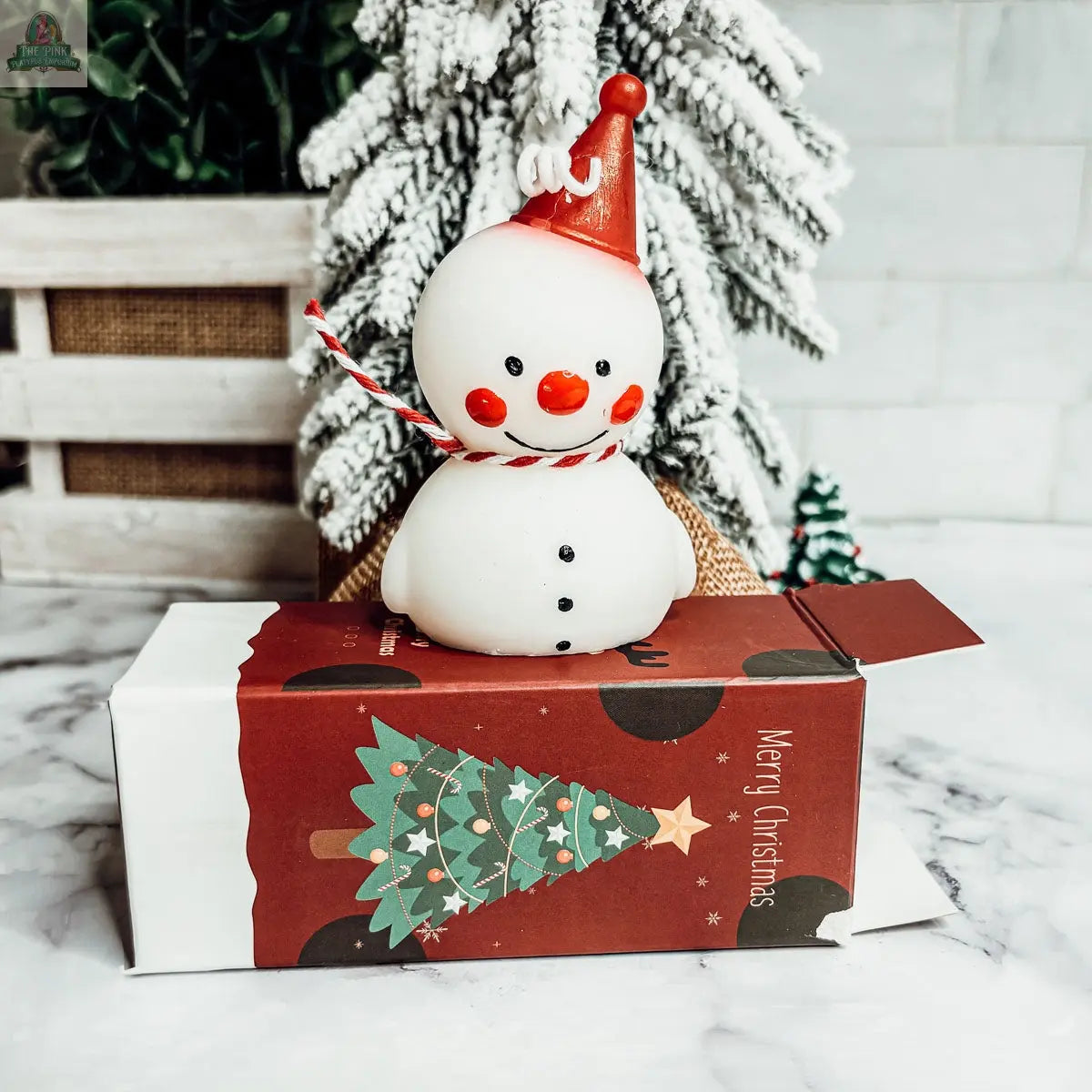 The Snowman Candle, featuring a red hat and scarf, sits atop a festive red Christmas box with an artificial snowy pine tree—perfect for winter decor or as a charming holiday gift.