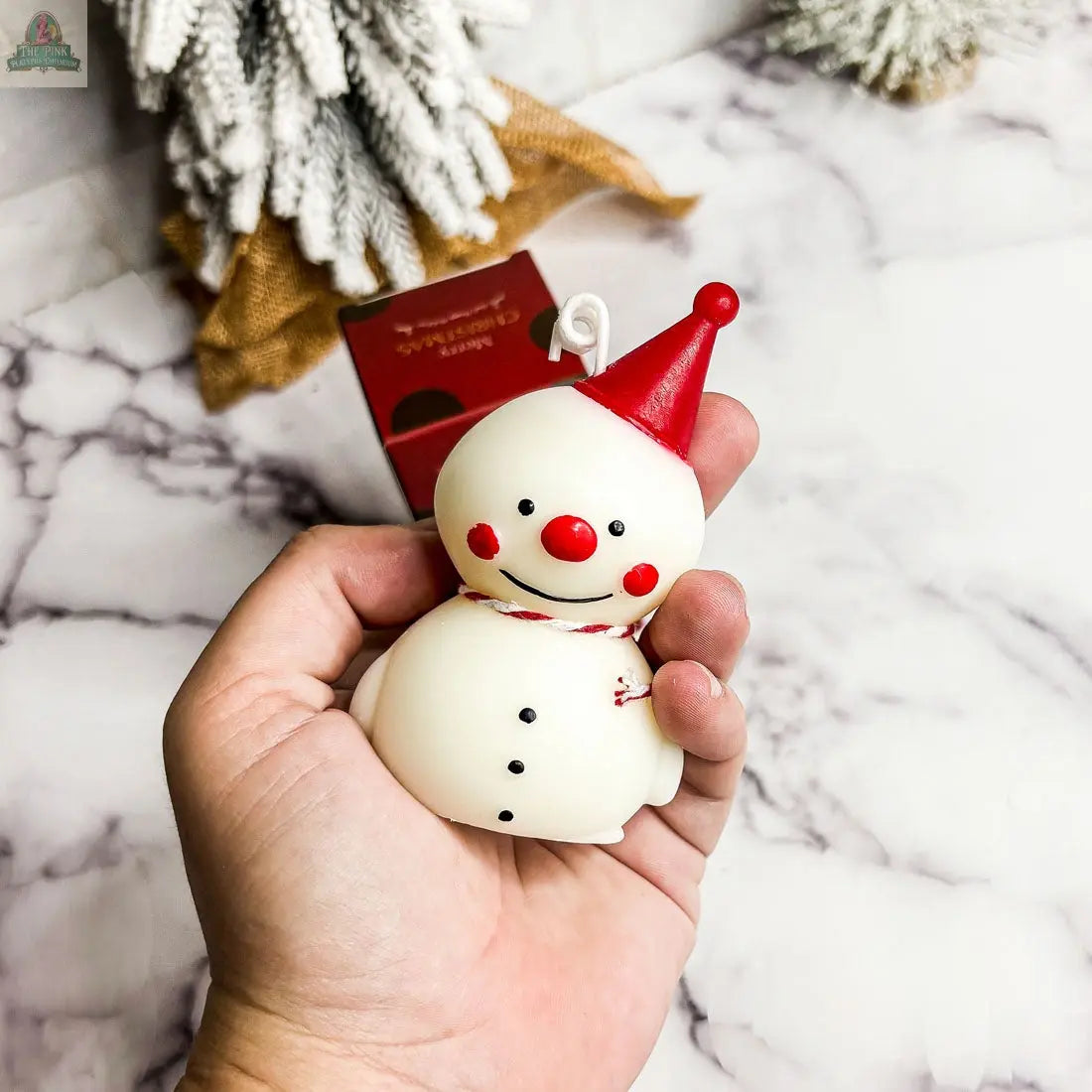 A hand holds the Snowman Candle, featuring a red hat, nose, and scarf—ideal for gifting or adding festive cheer to your winter decor. Blurred Christmas ornaments and a red box sit on a marble surface in the background.