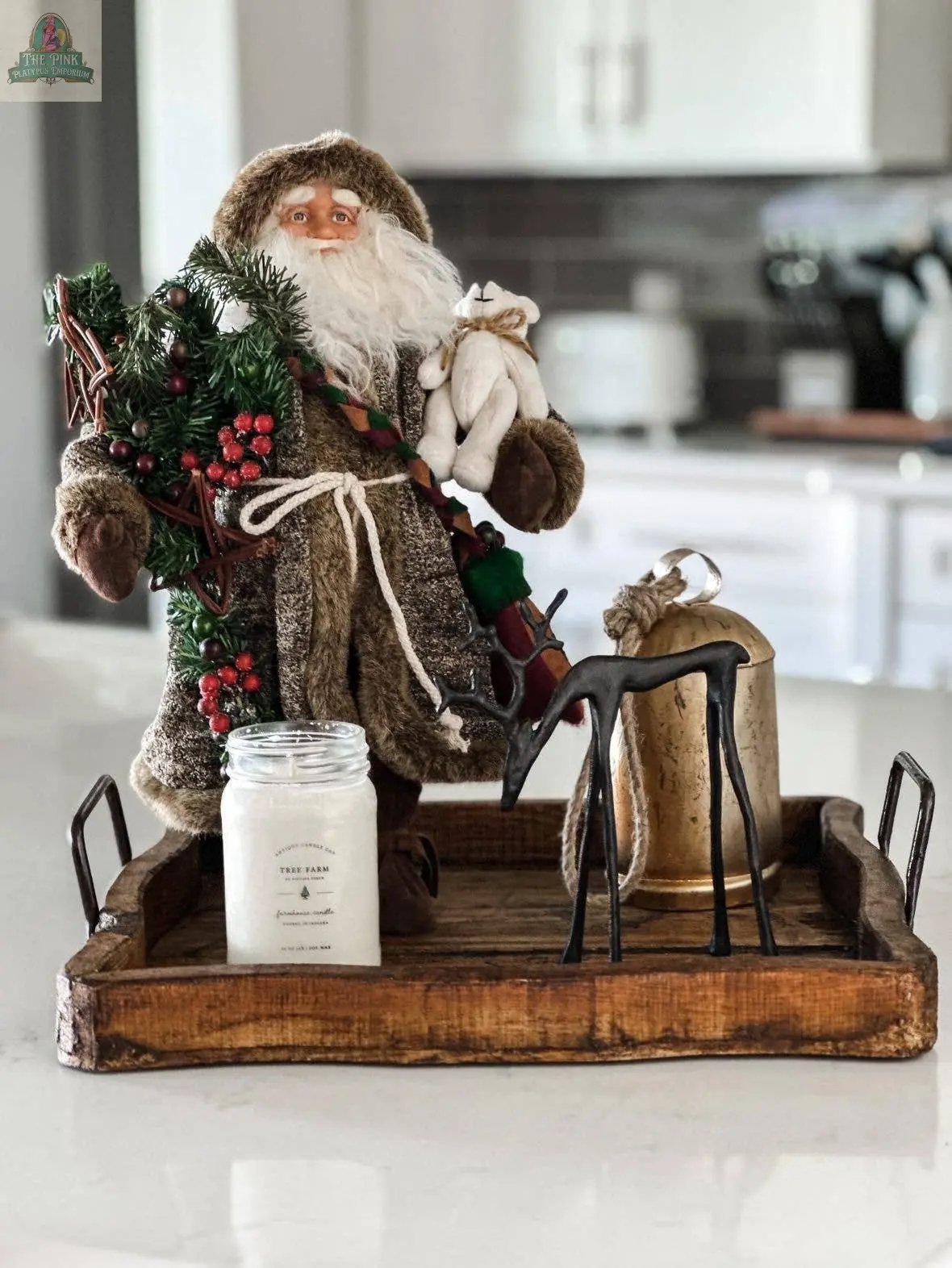 A decorative Santa with greenery and a rabbit stands on the handmade Natural Square Wood Tray (7.2"), alongside a candle, metal reindeer, and gold bell, atop a kitchen countertop.