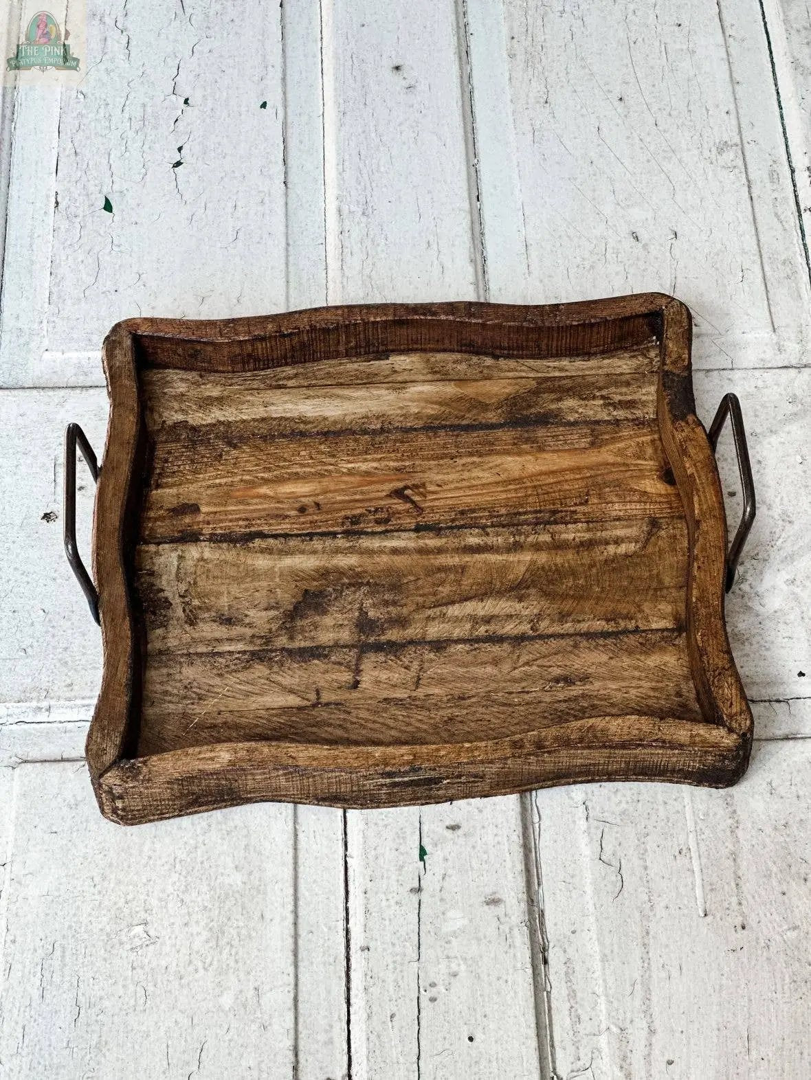 The handmade Square Wood Tray, Natural, 7.2, with a rustic, weathered finish and curved metal handles, rests on a distressed white wooden surface, highlighting its natural wood charm.