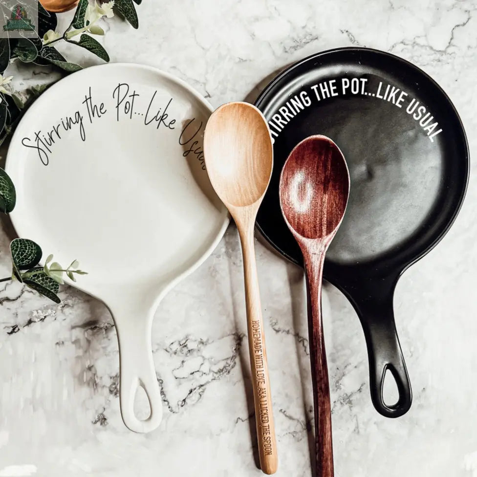 The "Stirring the Pot" set includes white and black porcelain spoon rests, each labeled "Stirring the pot...like usual" and holding a wooden spoon, displayed on marble—an ideal kitchen gift.