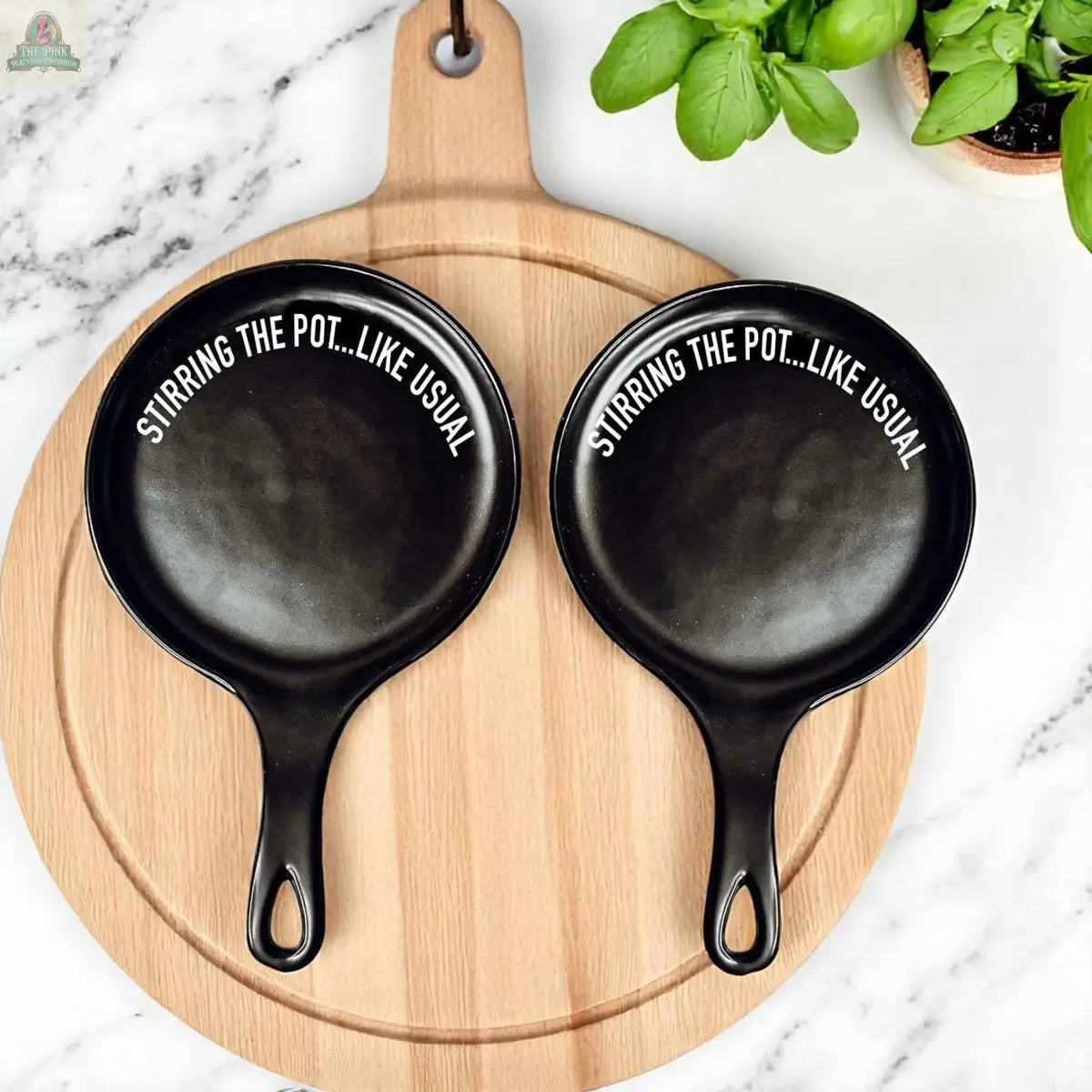 The "Stirring the Pot" set features two black pans on a wooden cutting board, paired with a stylish porcelain spoon rest—an elegant kitchen gift for any chef.