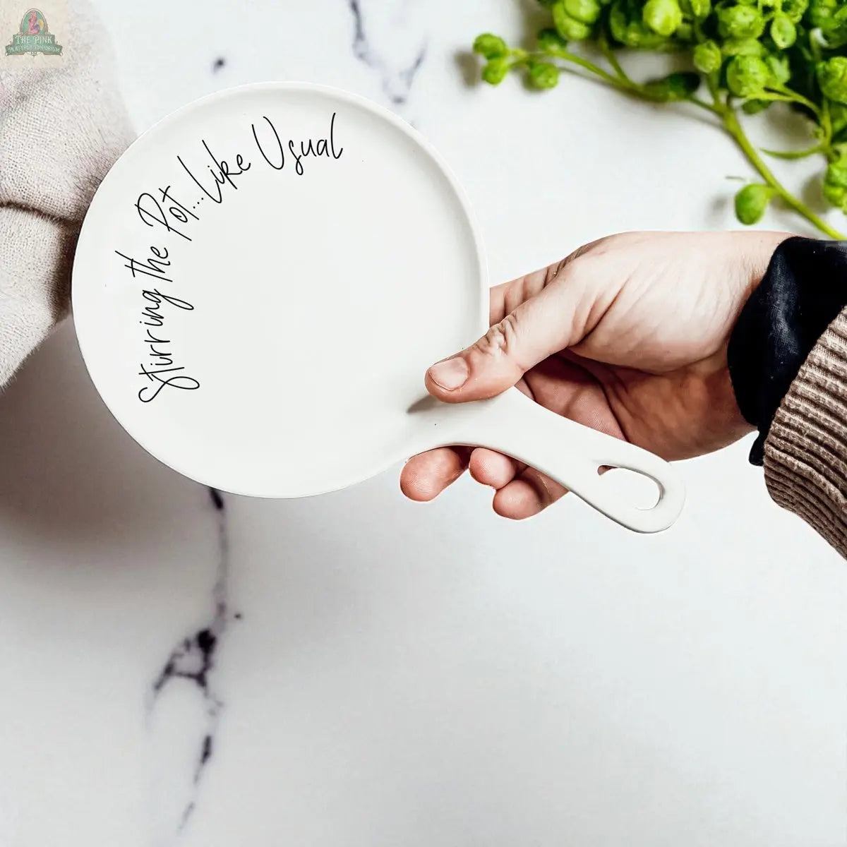 A hand holds the "Stirring the Pot" porcelain spoon rest, featuring its playful phrase; a green leafy plant and marble countertop are in the background—a charming kitchen gift for any cook.