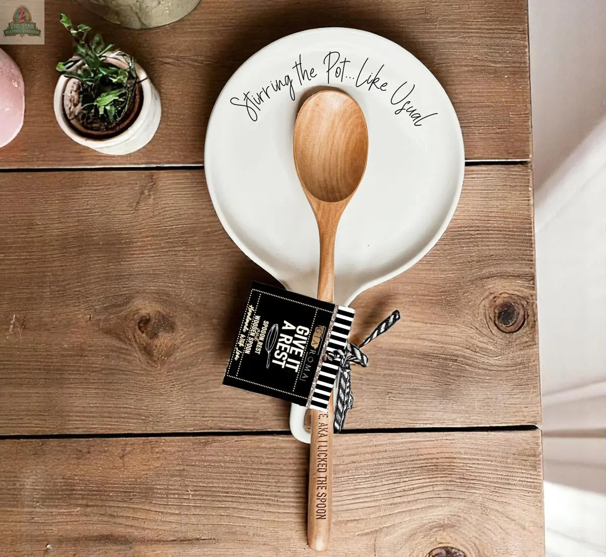 A wooden spoon named "Stirring the Pot" rests on a white plate, tied with a black-and-white "GIVE ME A REST" tag—an ideal kitchen gift displayed on a rustic wooden table.