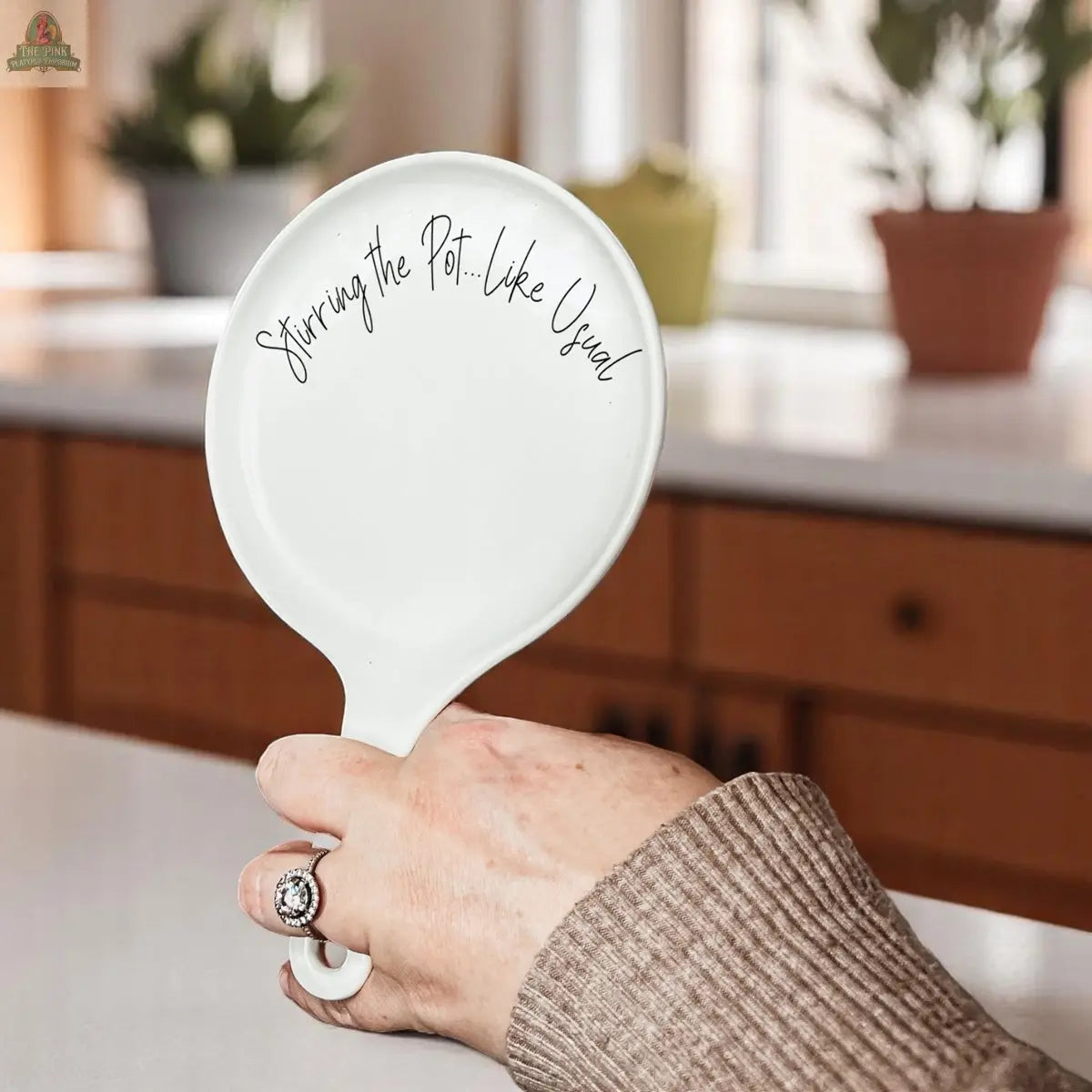A hand holds the "Stirring the Pot" porcelain spoon rest with “Stirring the pot... like usual” in black script—a charming kitchen gift. Warm wooden cabinets and potted plants complete the cozy background.
