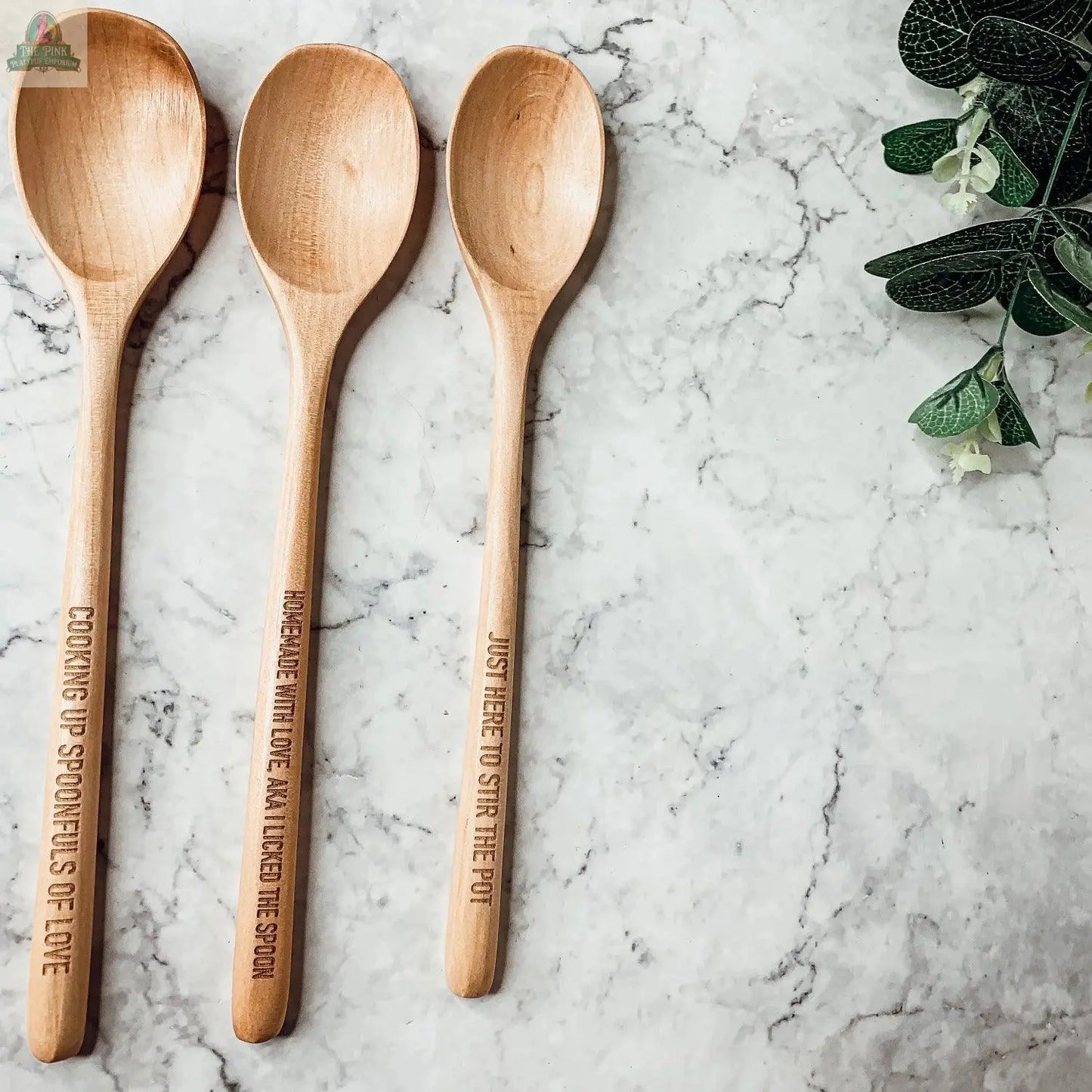 Three "Stirring the Pot" wooden spoons, each with a unique engraved phrase, rest on white marble beside green leaves—making this set a thoughtful and practical kitchen gift for any cook.