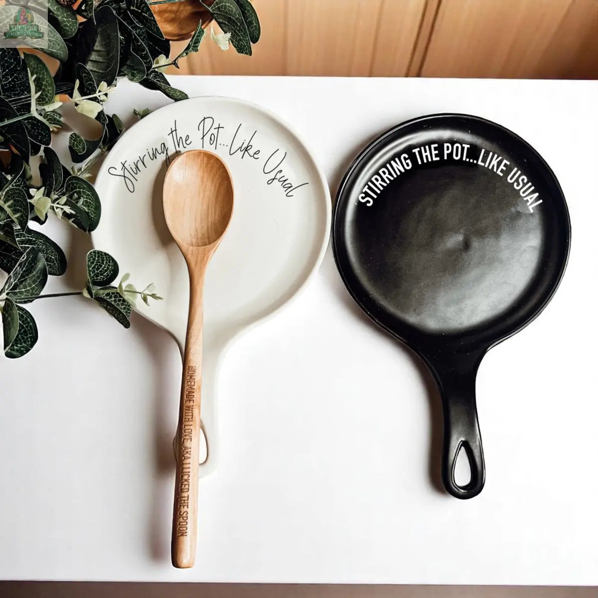 The "Stirring the Pot" set includes two porcelain spoon rests—one white, one black—each printed with "Stirring the Pot... Like Usual." The white rest holds a wooden spoon. A stylish kitchen gift; green leaves peek in from the left.