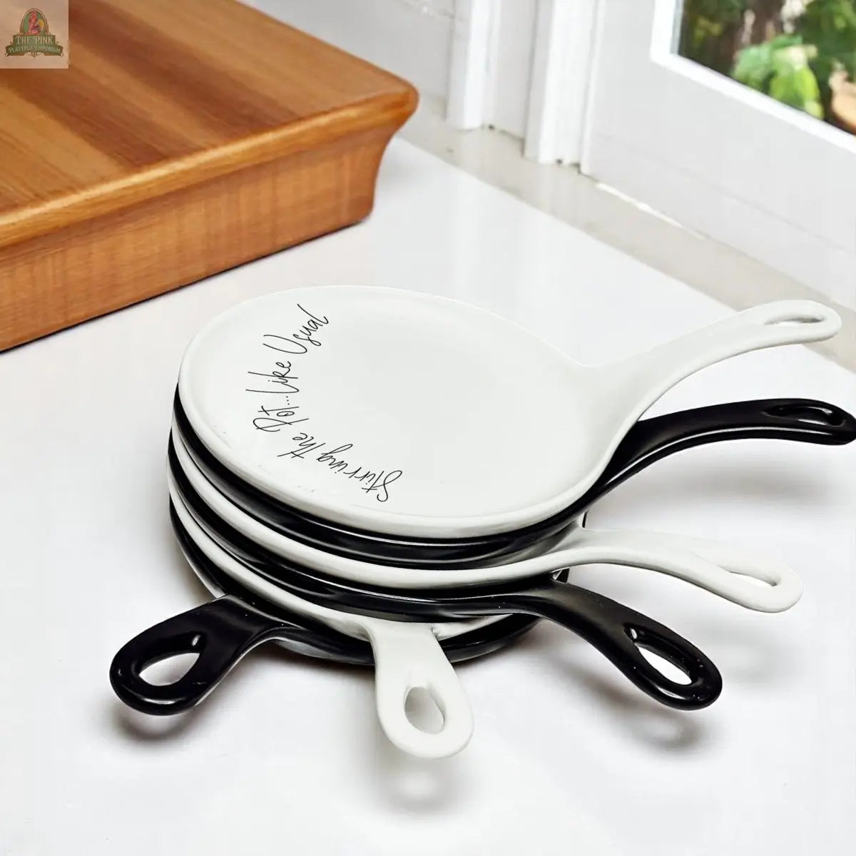 A stack of five white ceramic plates with black handles sits on a kitchen counter near a window. The top plate, named Stirring the Pot, features cursive writing and makes a perfect kitchen gift or porcelain spoon rest.