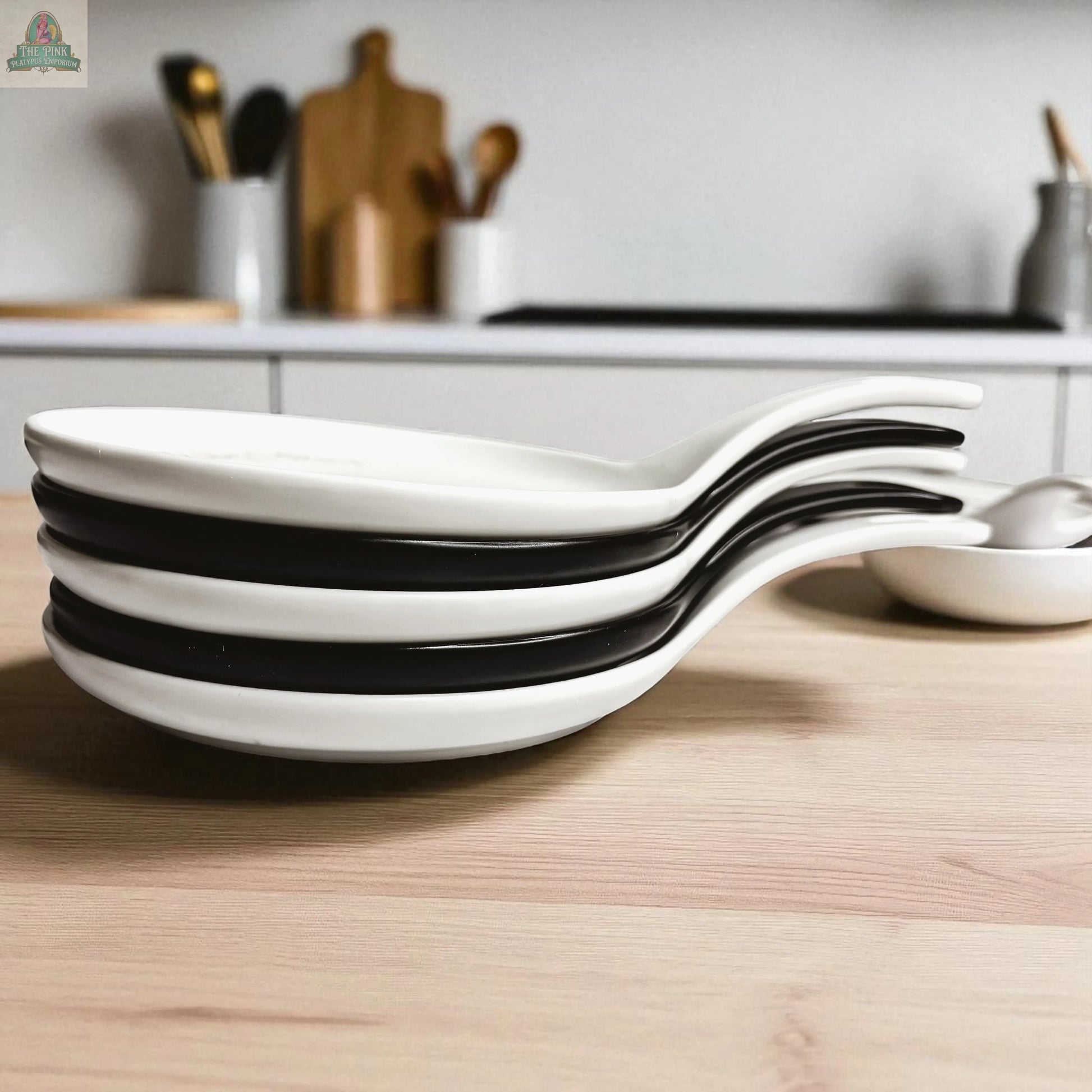 Stirring the Pot: Four white ceramic soup spoons with black accents are stacked on a wooden countertop beside a porcelain spoon rest, making an ideal kitchen gift in a modern setting with blurred utensils and cutting boards behind.