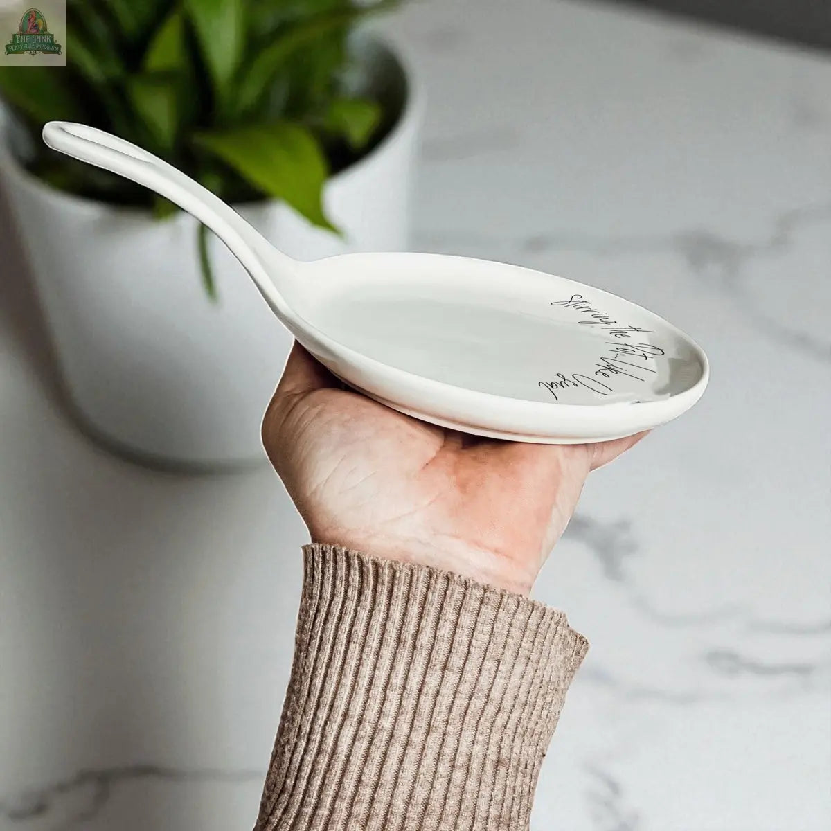 A person holds the "Stirring the Pot" porcelain spoon rest with a handle and elegant cursive writing. A green potted plant on a white marble countertop adds to its charm, making it a perfect kitchen gift.