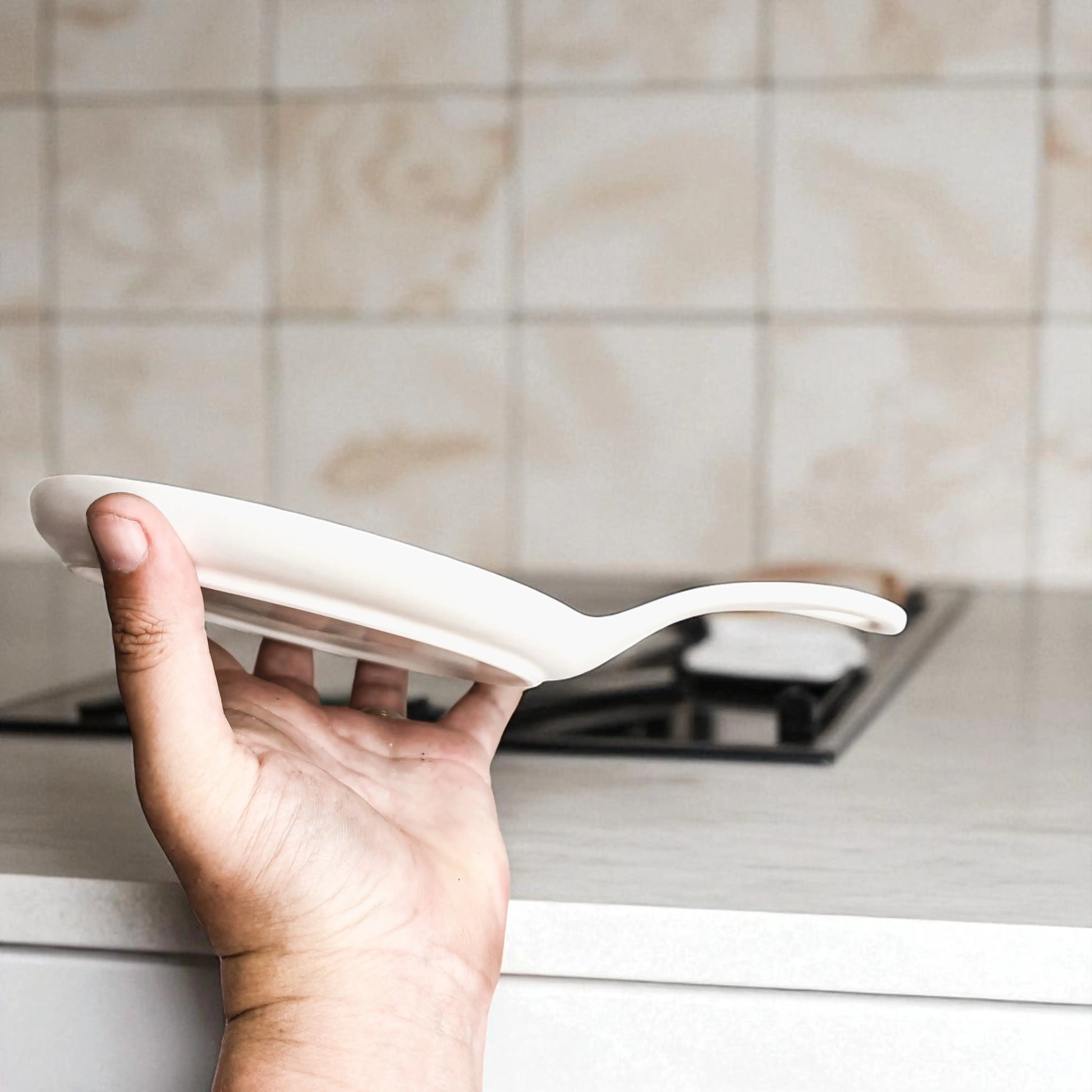 A hand holds the "Stirring the Pot" white ceramic frying pan above a light kitchen counter next to a porcelain spoon rest, with a gas stove and tiled backsplash in the background—the perfect kitchen gift.