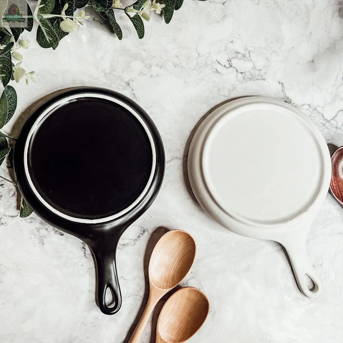 Stirring the Pot" ceramic skillet-shaped plates, ideal as spoon rests or distinctive kitchen gifts, are displayed upside down on marble with two wooden spoons and fresh green leaves enhancing the charming setup.