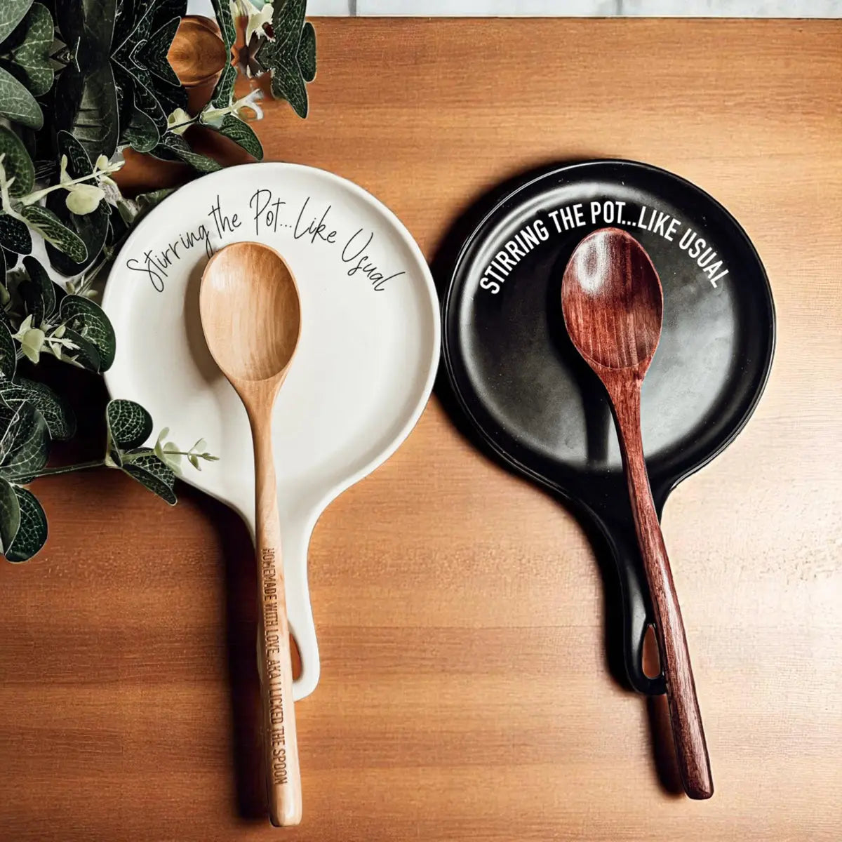 The "Stirring the Pot" set includes black and white ceramic spoon rests, each with a wooden spoon. Featuring green leaf designs and the phrase "Stirring the Pot... Like Usual," it makes a funny kitchen gift.