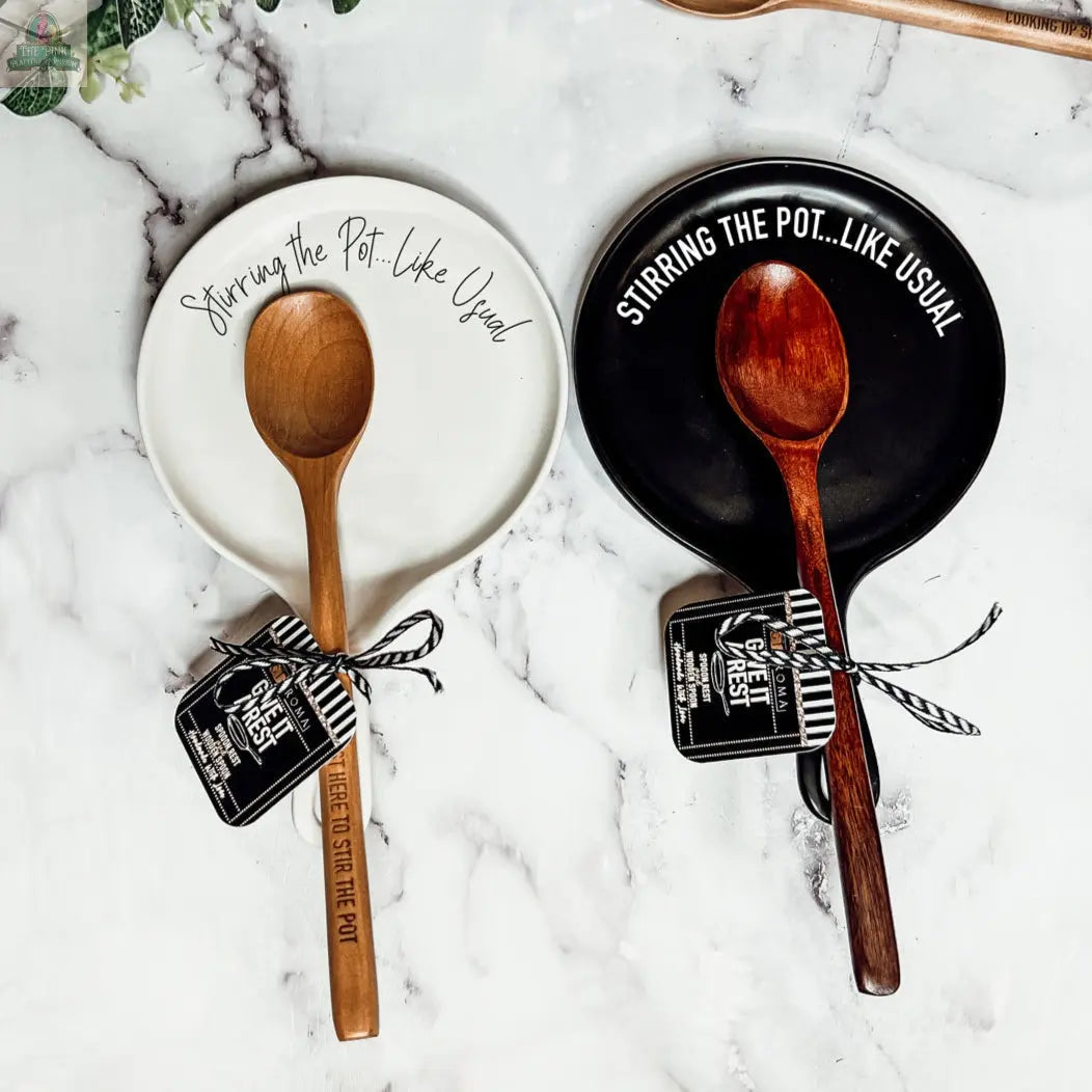 The "Stirring the Pot" set features two porcelain spoon rests (one white, one black), each with a wooden spoon and the phrase Stirring the Pot... Like Usual—an ideal kitchen gift displayed on a white marble surface.