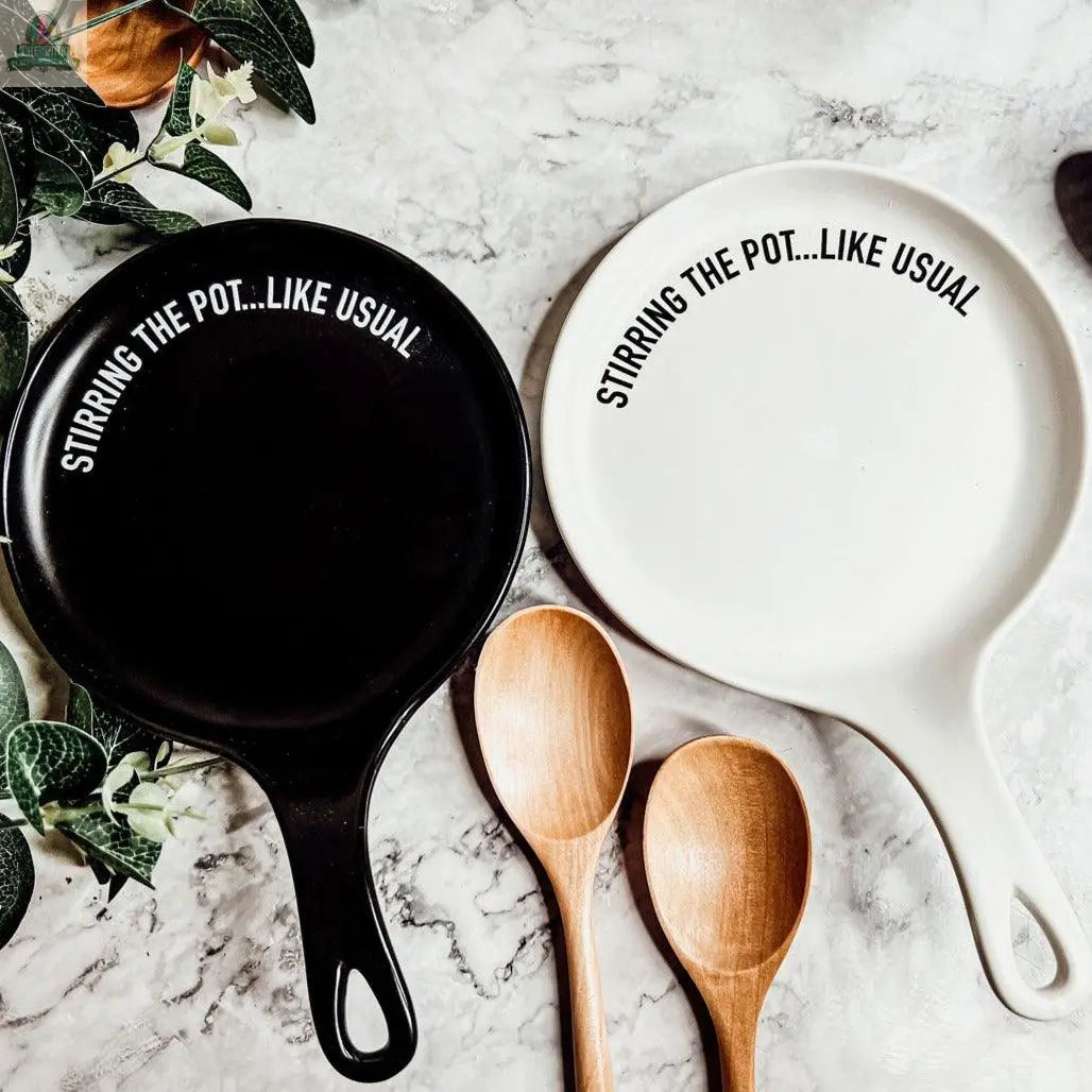 The Stirring the Pot set includes two porcelain spoon rests, one black and one white, each labeled “STIRRING THE POT...LIKE USUAL.” Displayed on marble with wooden spoons and green leaves, it makes a humorous kitchen gift!.