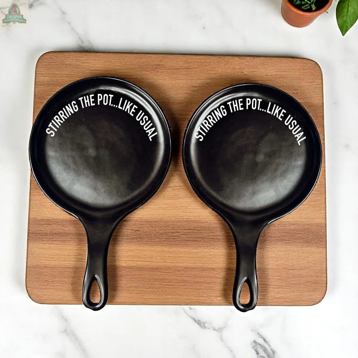 Two black "Stirring the Pot" cast iron skillets sit side by side on a wooden cutting board atop a marble countertop, with a small potted plant and porcelain spoon rest nearby.
