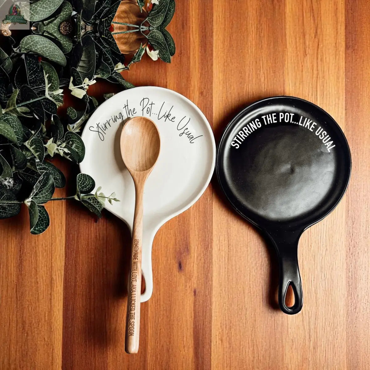 Two "Stirring the Pot" spoon rests—one white with a wooden spoon, one black—sit on a wooden surface beside a plant. These porcelain kitchen accessories make a funny and stylish gift.