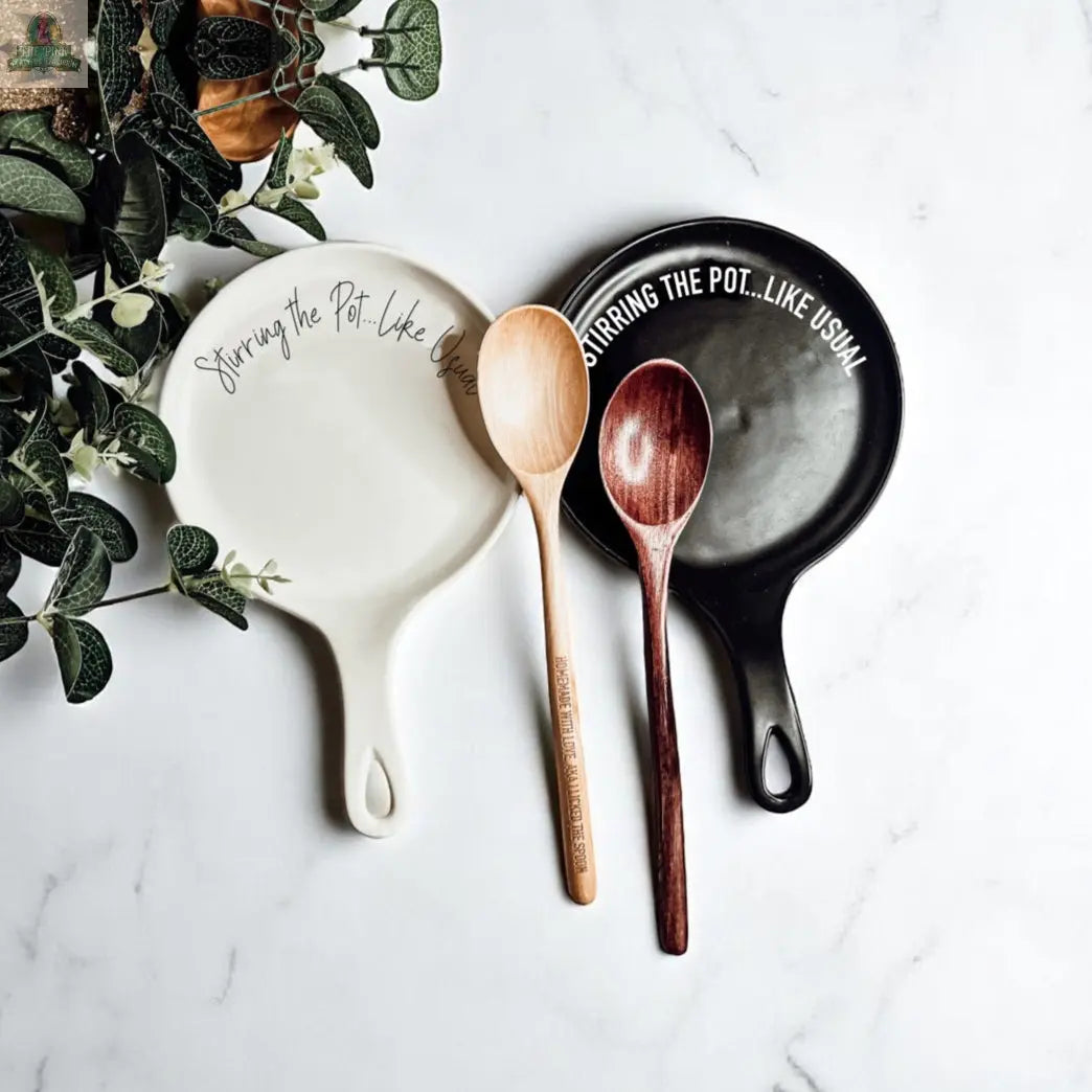 The “Stirring the Pot” set features two porcelain spoon rests—one black, one white—each with “Stirring the Pot…Like Usual,” shown with wooden spoons and leafy greens. A fun kitchen gift perfect for any cook's counter.