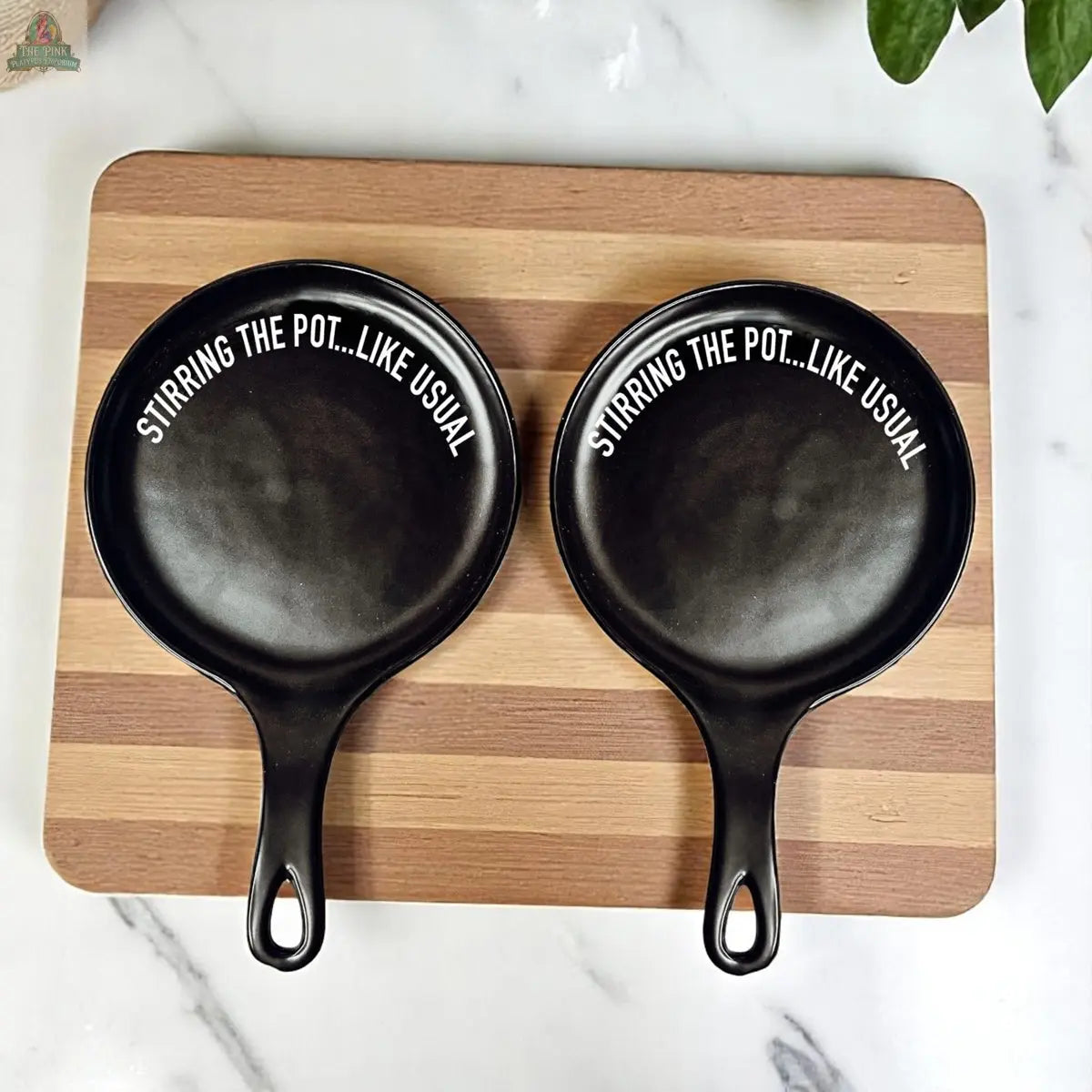 Two small black Stirring the Pot pans with handles sit side by side on a striped wooden cutting board. Each witty porcelain kitchen accessory features “STIRRING THE POT...LIKE USUAL” in white text on the inside.