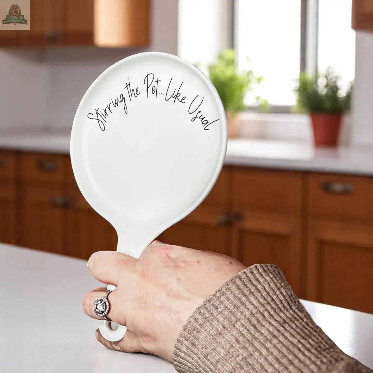 A hand holds the "Stirring the Pot" white pan-shaped spatula in a kitchen with wooden cabinets and potted plants—a quirky porcelain kitchen accessory and a perfect funny gift.