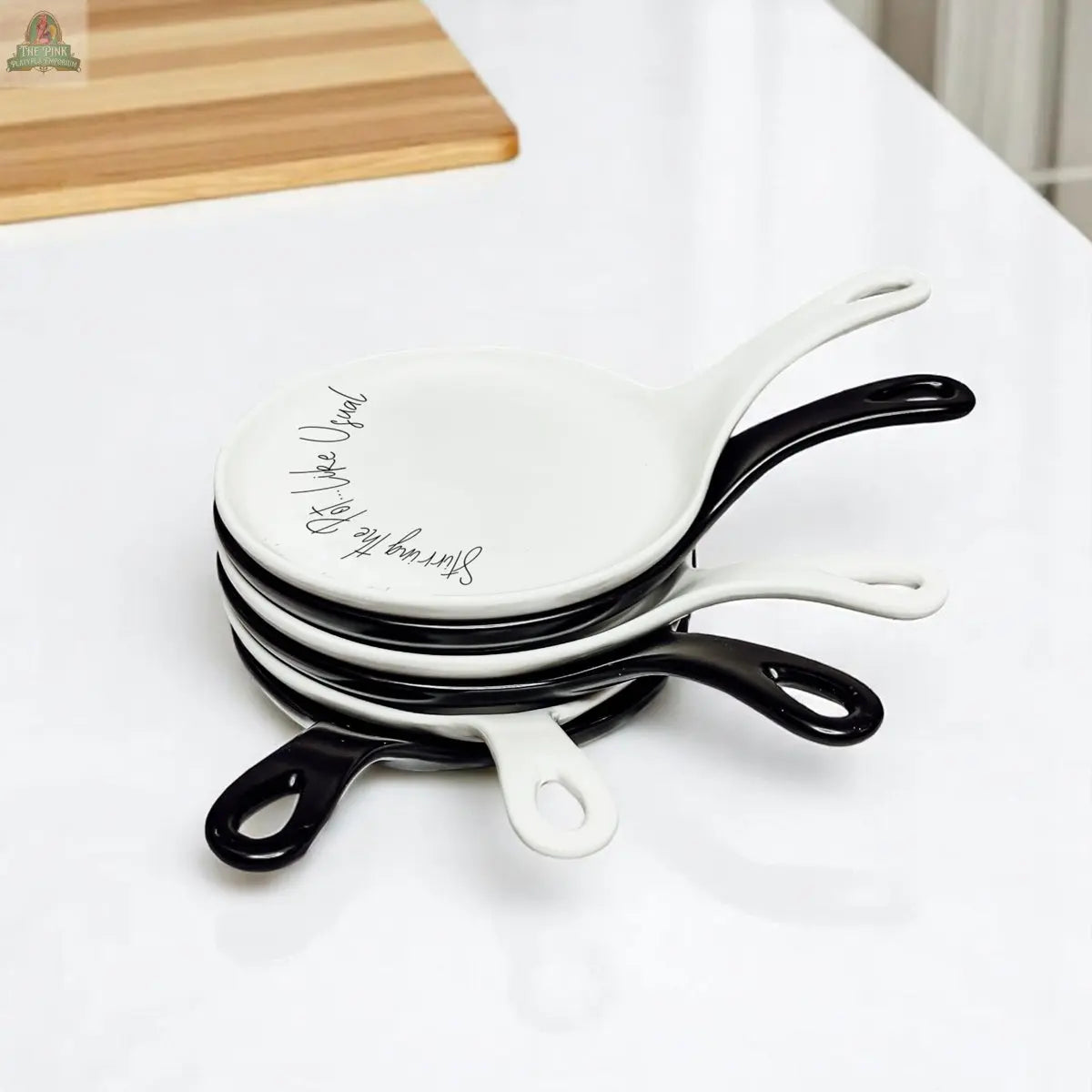 A stack of four small Stirring the Pot porcelain plates with handles, shaped like mini skillets, sits on a white surface. The top plate is white with cursive writing; the rest alternate black and white.