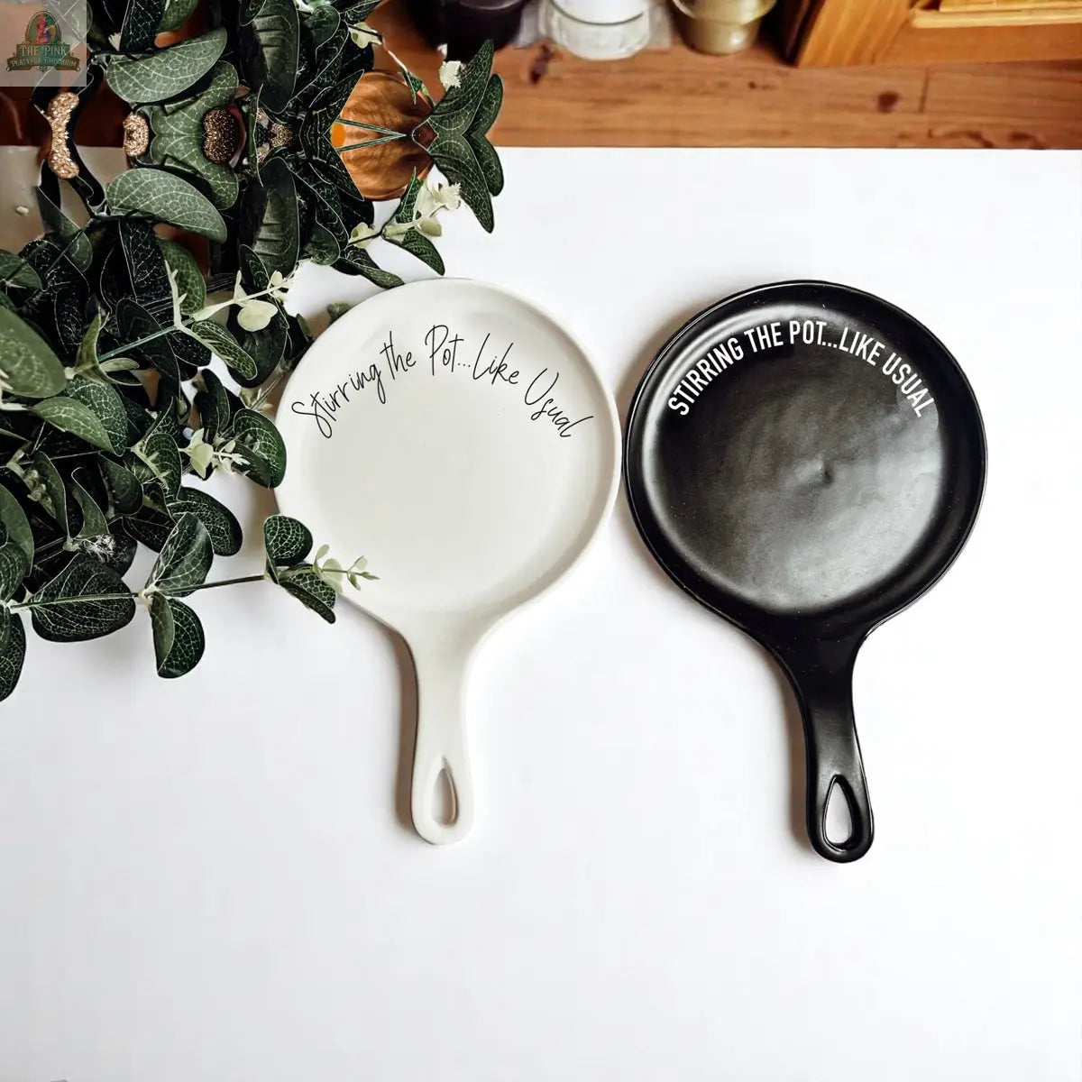 Two "Stirring the Pot" porcelain spoon rests, one white and one black, are displayed side by side on a white surface near a leafy plant—making them the perfect kitchen gift for any home cook.