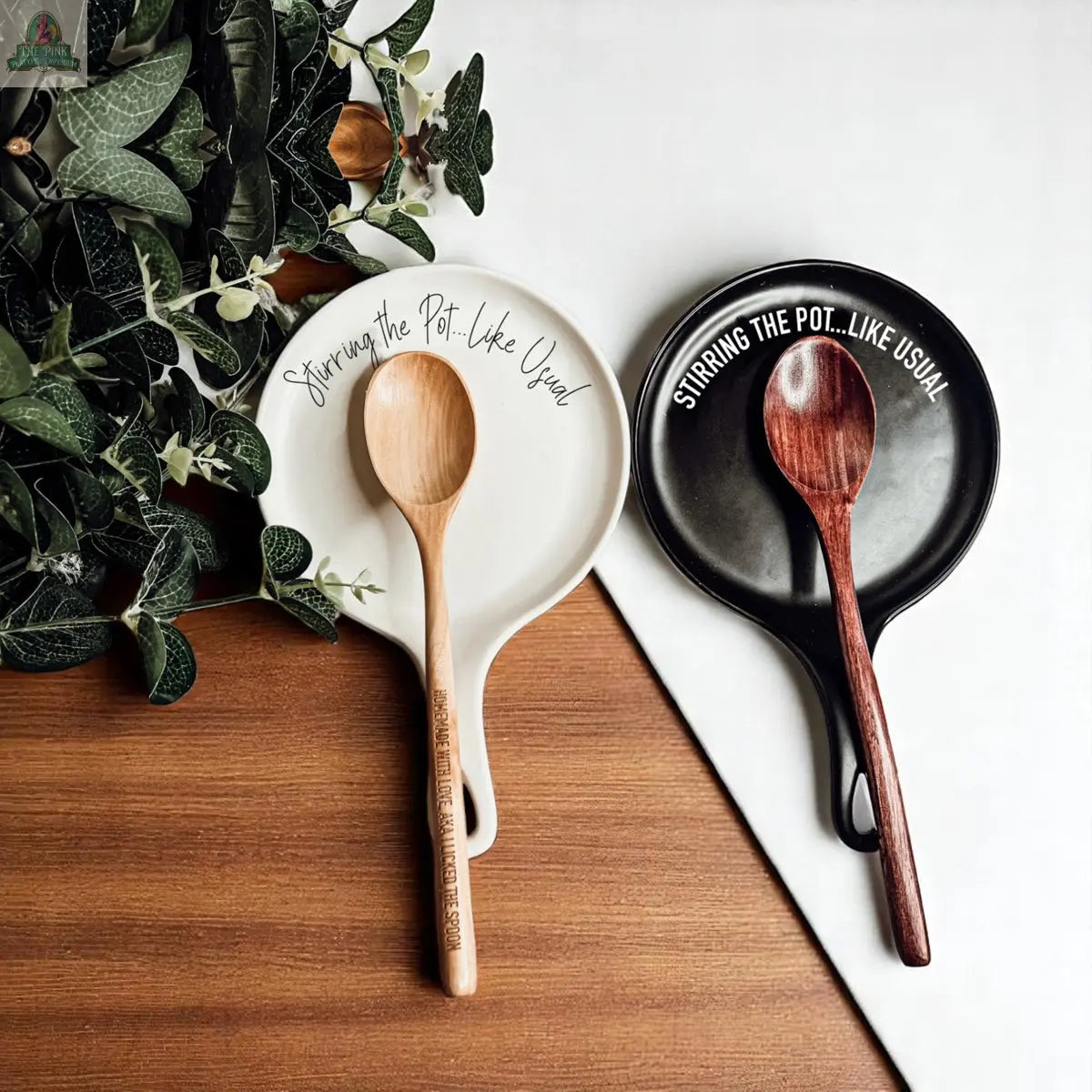 Two wooden spoons labeled “Stirring the Pot...Like Usual” rest on a black and white porcelain holder. Green leaves add charm, making the “Stirring the Pot” set a perfect kitchen gift on a wood and white backdrop.