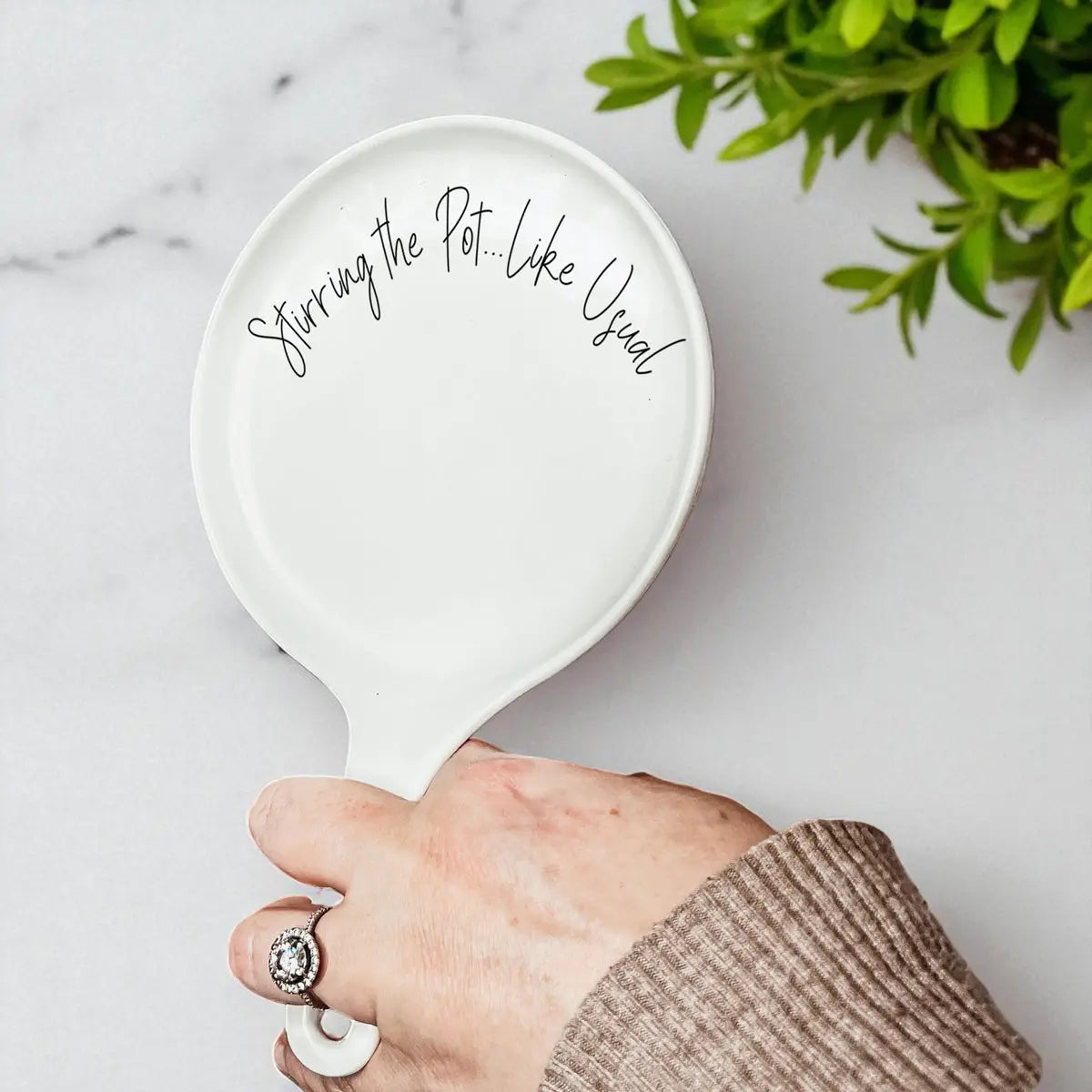 A hand holds the "Stirring the Pot" porcelain spoon rest beside a green potted plant on marble—a playful kitchen gift for any cook.