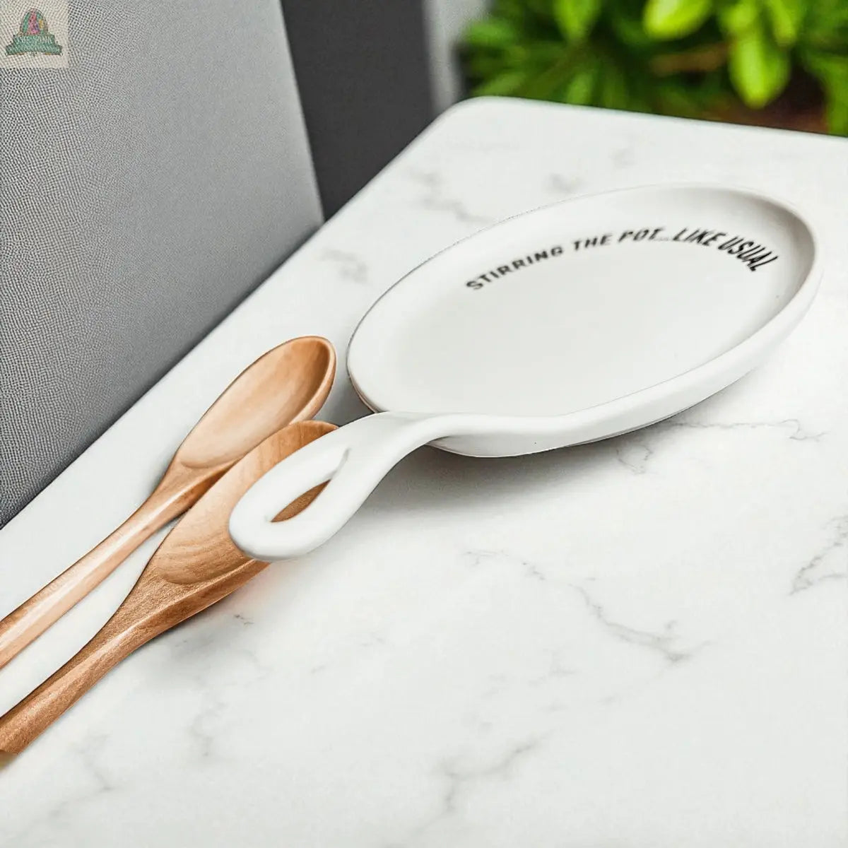 The Stirring the Pot white porcelain spoon rest, featuring the phrase "STIRRING THE POT, LIKE USUAL," sits beside two wooden spoons on a marble surface with blurred green leaves in the background—a unique kitchen gift.