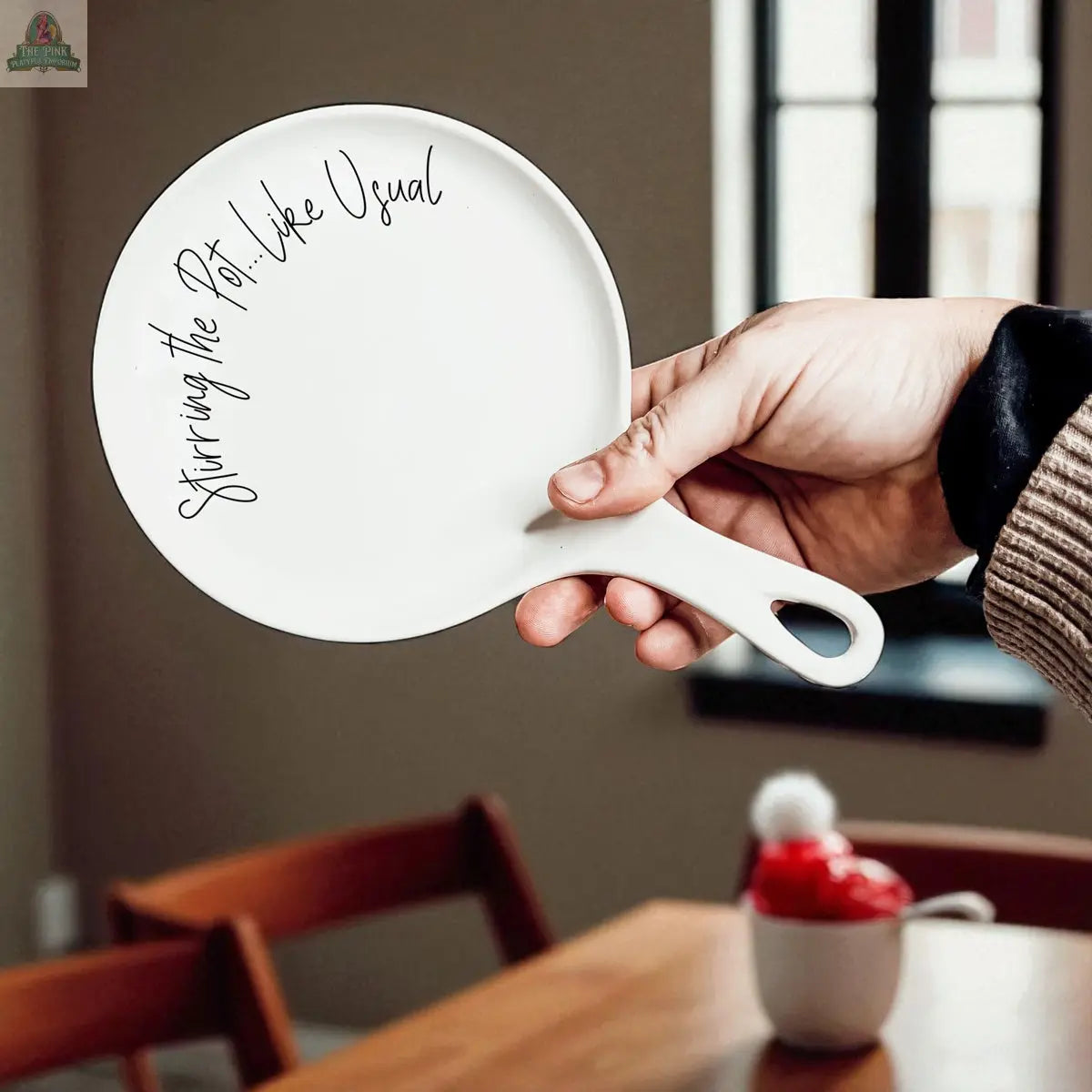 A hand holds the “Stirring the Pot” white porcelain spoon rest, shaped like a pan and featuring black script—a charming kitchen gift seen against a wooden table and sunlit window.