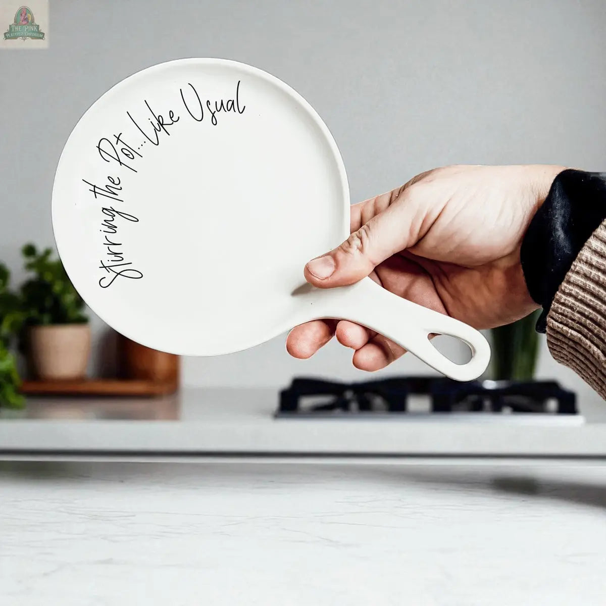 A hand holds the Stirring the Pot white porcelain spoon rest, shaped like a pan and inscribed with "Stirring the Pot. Like Usual." A stove and potted plants appear in the background—an ideal kitchen gift for any home cook.