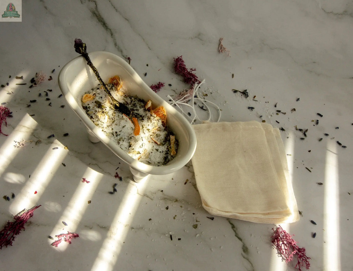 A small white bathtub dish holds Stress Relief dried flowers, herbs, and Epsom salt bath mix with a spoon. Folded cloth bags rest nearby on marble with scattered petals—perfect for a calming herbal bath tea ritual.