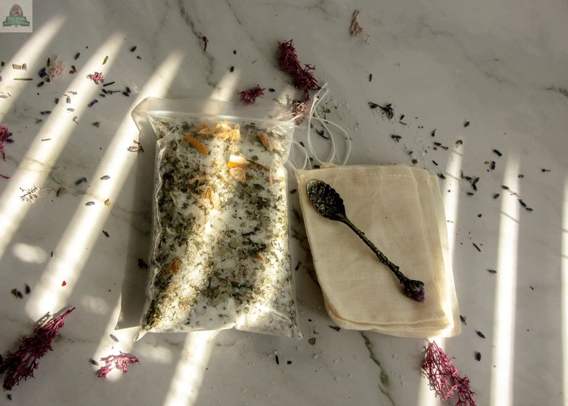 A packet of Stress Relief dried herbs for an herbal bath tea rests on a marble surface beside stacked muslin bags and an ornate metal spoon, with dried purple flowers and scattered herbs in patches of sunlight.