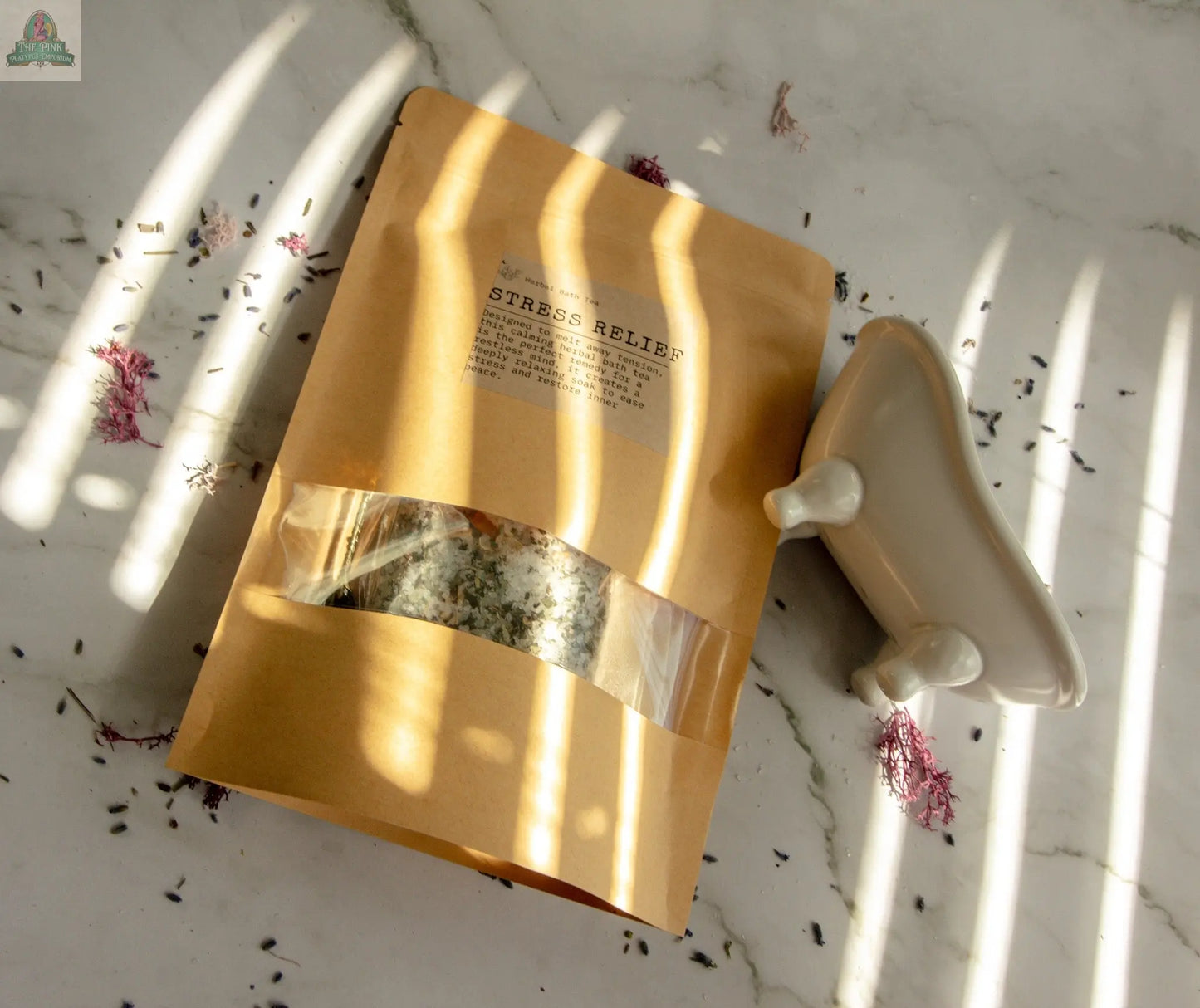 A brown pouch labeled Stress Relief bath soak rests on a marble surface with dried flowers. Nearby, a white clawfoot tub dish sits as sunlight casts striped shadows, creating a calming scene perfect for relaxation.