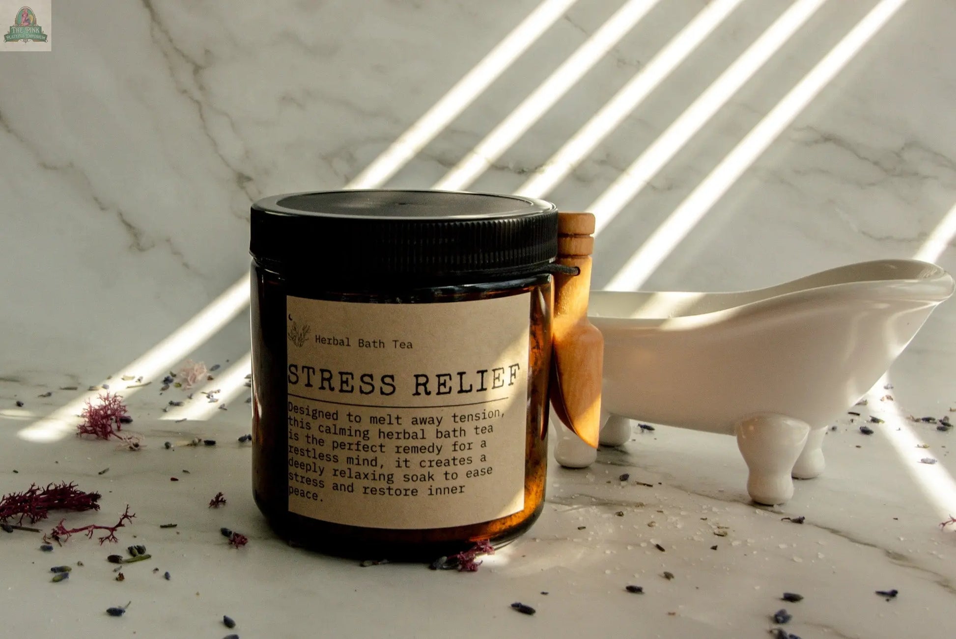 A jar of Stress Relief bath tea rests on marble with dried herbs around it. Sunlight filters over a small white clawfoot tub and wooden scoop, hinting at a soothing Epsom salt bath ritual.