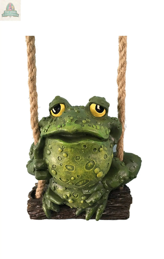 A green, bumpy frog with yellow eyes sits on a wooden swing with thick rope handles, looking upward. The whimsical TOAD SWINGER makes a charming addition to your outdoor decor. Background is plain white.