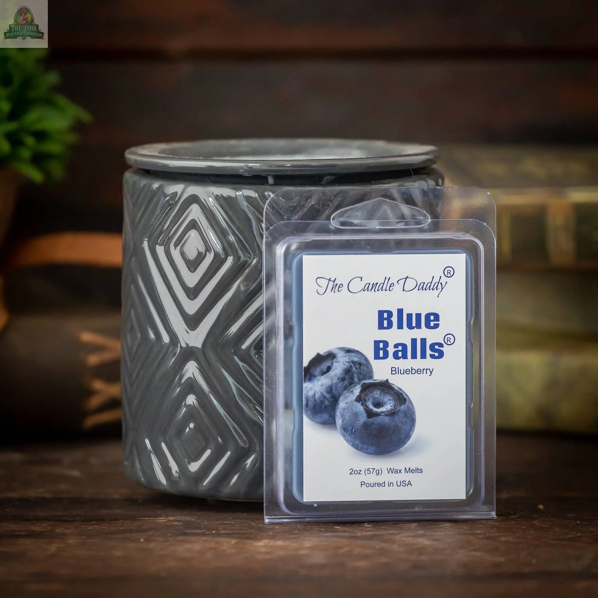 A gray patterned wax warmer sits behind a package of The Candle Daddy’s TWO BLUE BALLS - RIPE BLUEBERRY SCENTED WAX MELT, offering blueberry aromatherapy. Blurred books and a plant are visible in the background.