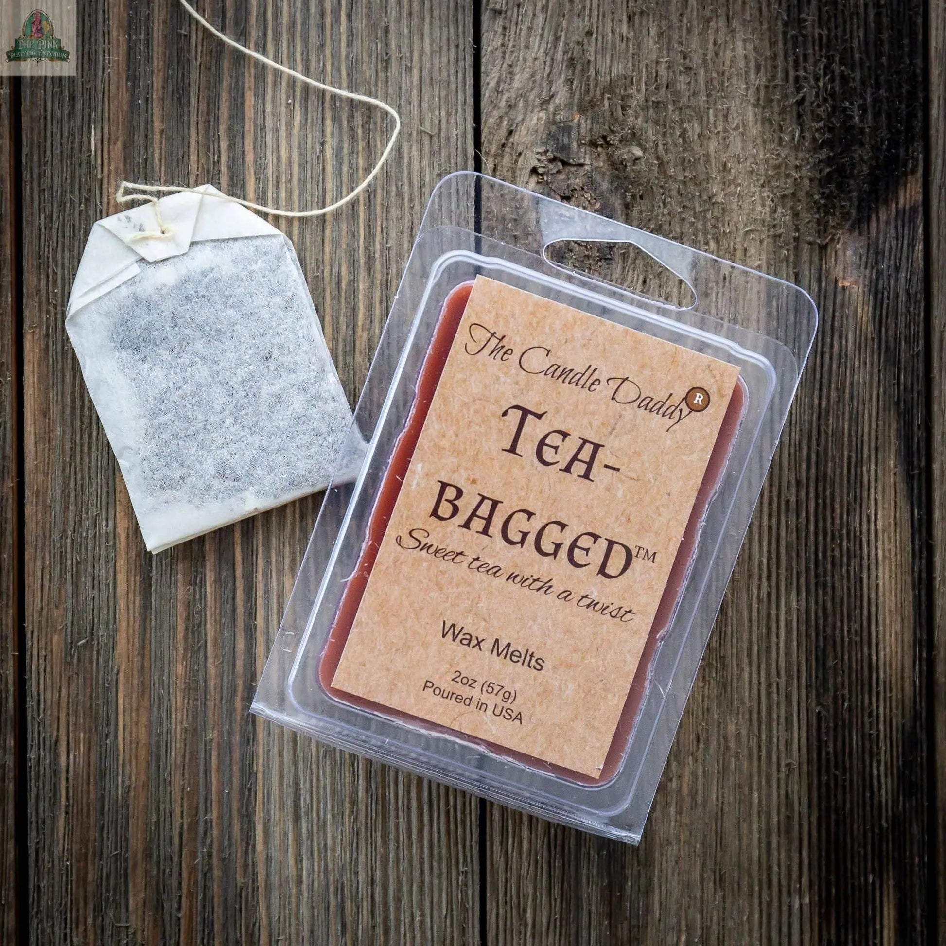 A tea bag lies beside a pack of “Tea-Bagged - Sweet Tea With A Twist Scented Melt- Maximum” wax melts by The Candle Daddy on a rustic wooden surface. The 2oz label notes it’s poured in the USA.