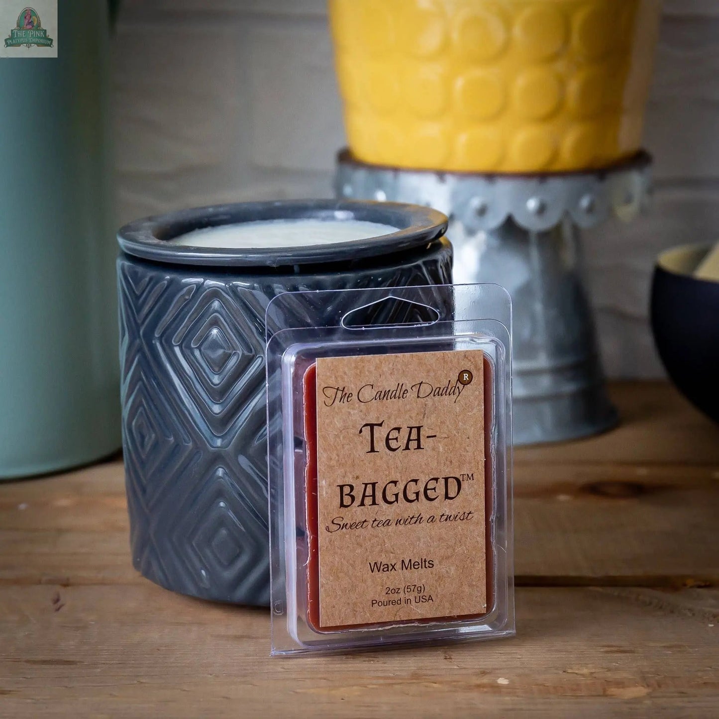 A package of Tea-Bagged - Sweet Tea With A Twist Scented Melt- Maximum sits before a dark gray wax warmer on a wooden surface, with a yellow planter and assorted items visible in the background.