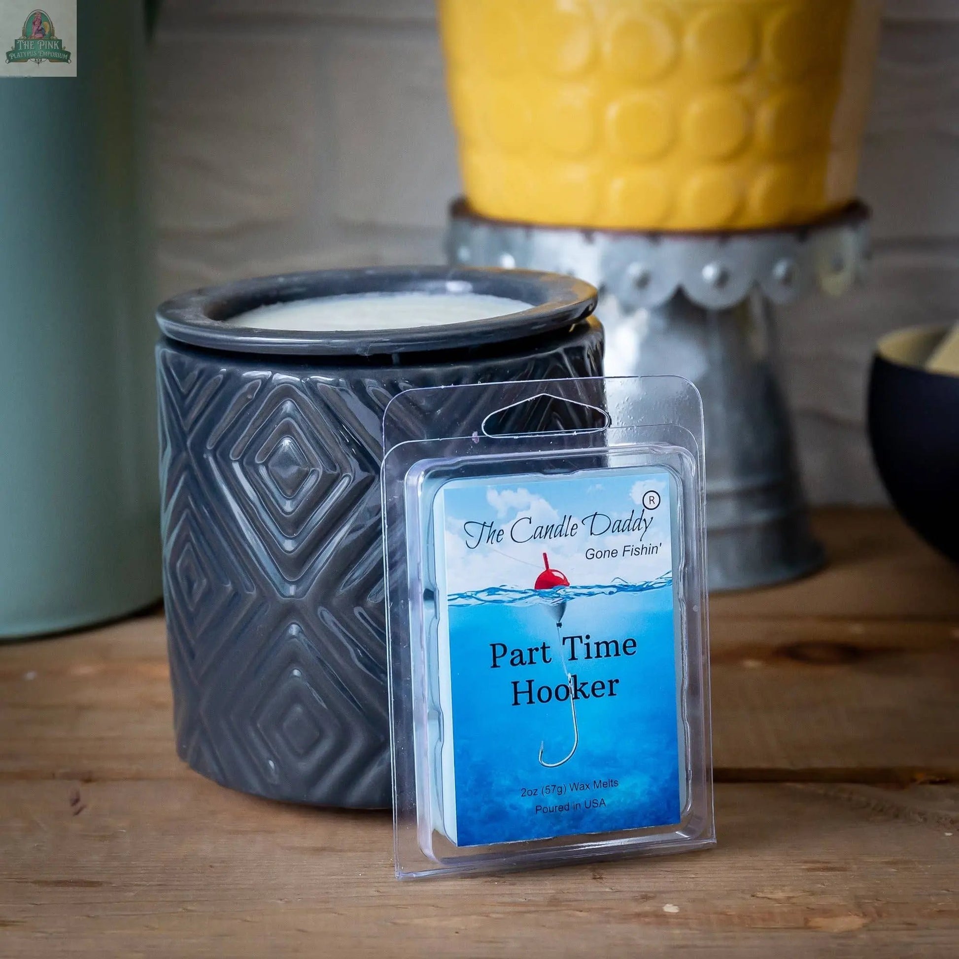 A gray patterned candle warmer sits on a wooden surface beside a pack of The Candle Daddy's Gone Fishin' - Part Time Hooker - Water's wax melts, ideal for aromatherapy. A yellow planter brightens the background.
