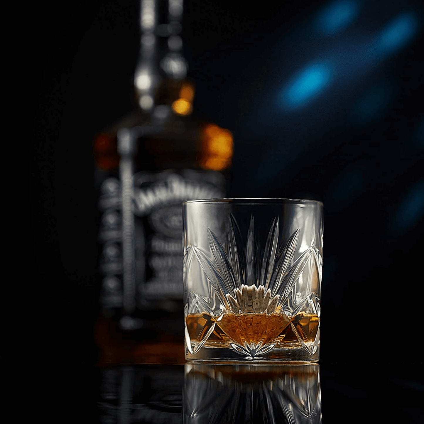 A cut crystal glass from The Connoisseur's Set - Palm Whiskey Glass Edition, filled with whiskey, sits in focus on a reflective surface, with a blurred bottle of Jack Daniels in the background against a dark backdrop.